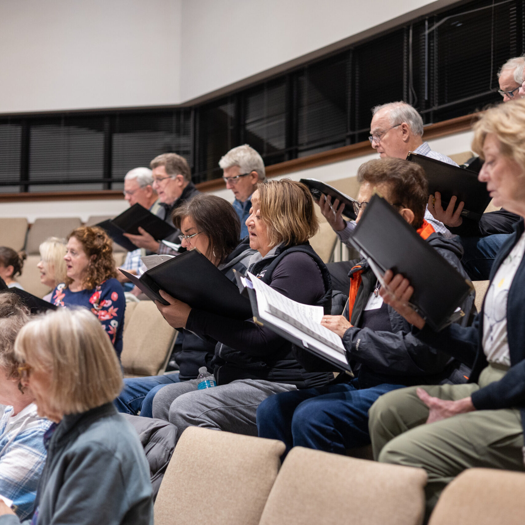 A group of adults sits on tiered seats, holding open music folders and singing together, appearing to participate in a choir rehearsal inside a well-lit room.