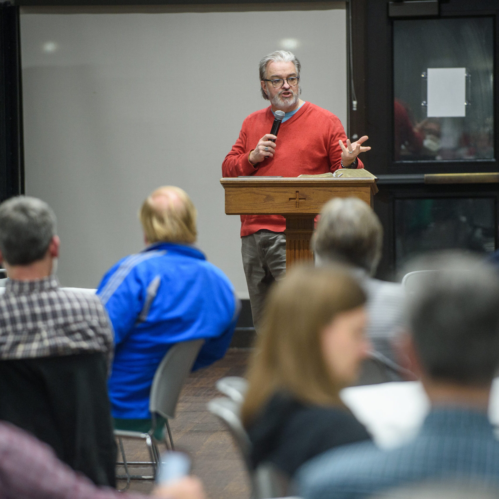 A man in a red sweater speaks into a microphone at a podium in front of a seated audience in a conference room.
