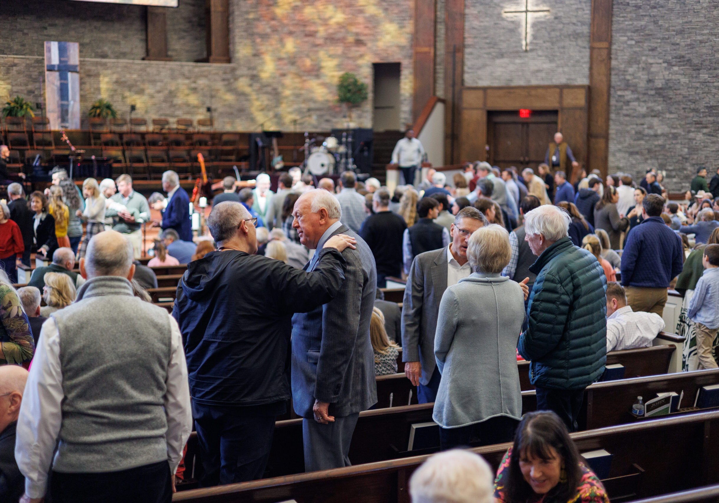 A large group of people gather inside a church, standing and talking in small groups. Some are shaking hands and greeting each other. The setting is spacious with rows of pews and a cross on the wall in the background.