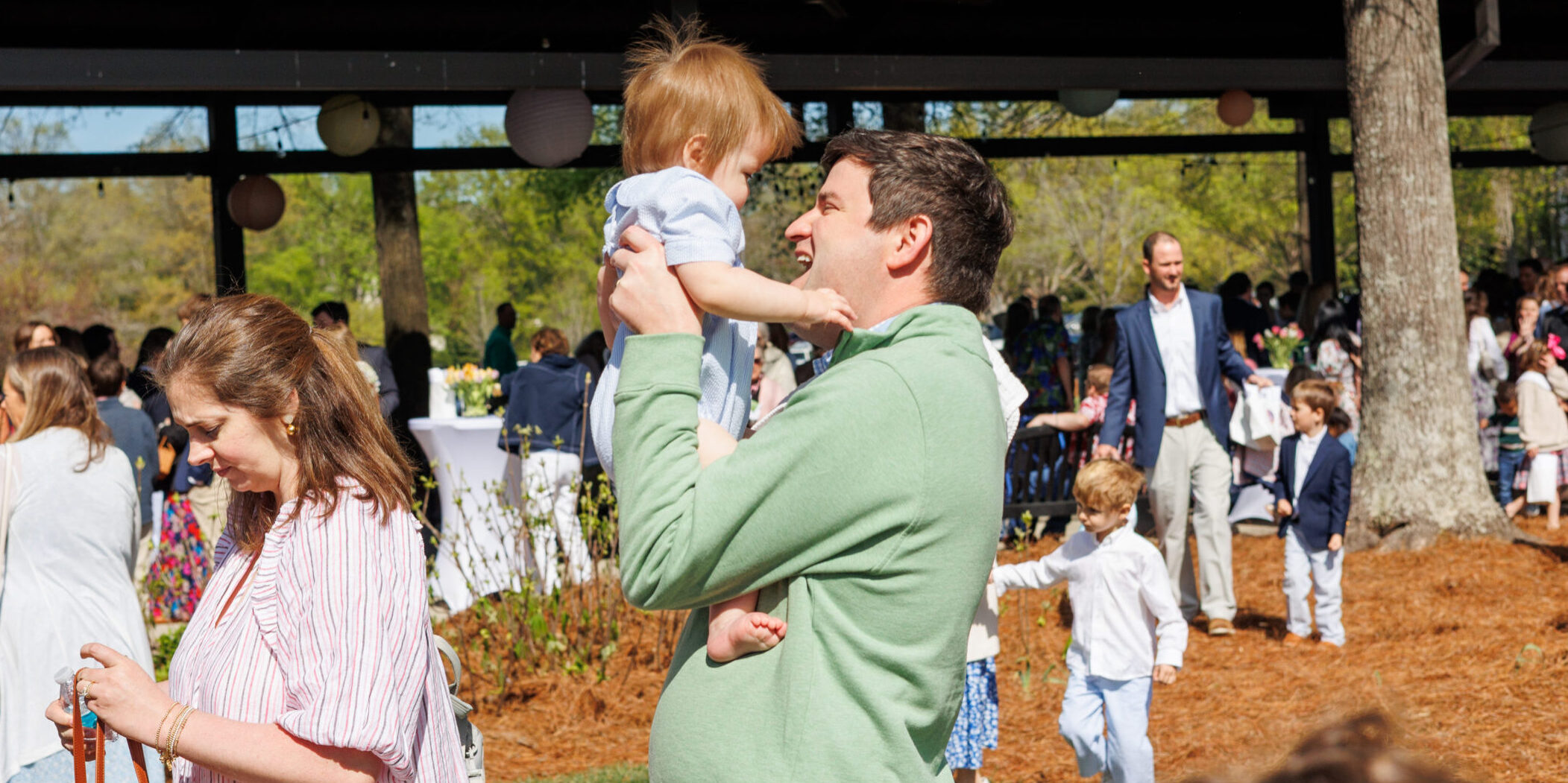 A man in a green sweater lifts a child in his arms at an outdoor gathering with many people, including children, near a pavilion on a sunny day. Trees and decorations are visible in the background.