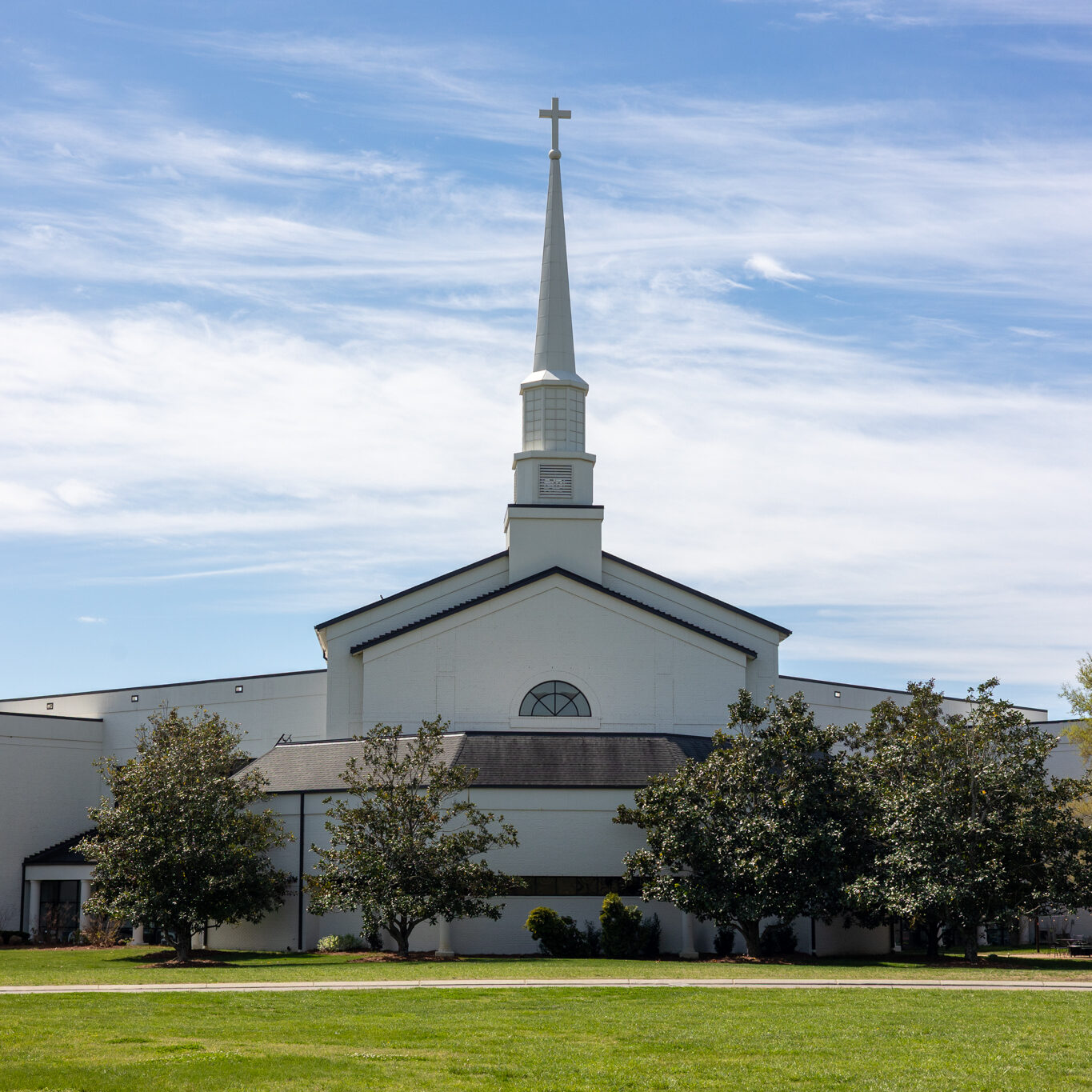 A white church with a tall steeple and cross stands on a grassy lawn under a blue sky with wispy clouds. A white car is parked on the left side near the building. Trees line the front of the church.