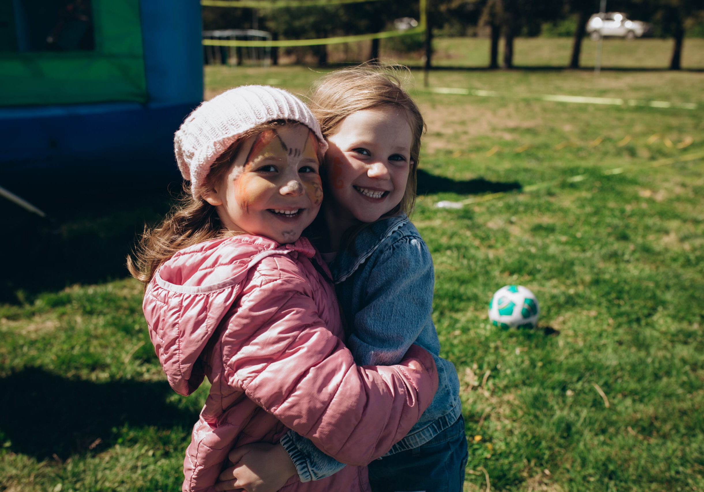 Two young girls smiling and hugging outdoors on grass, one wearing a pink jacket and hat with face paint, the other in a denim jacket. A green and blue soccer ball and inflatable play structure are in the background.