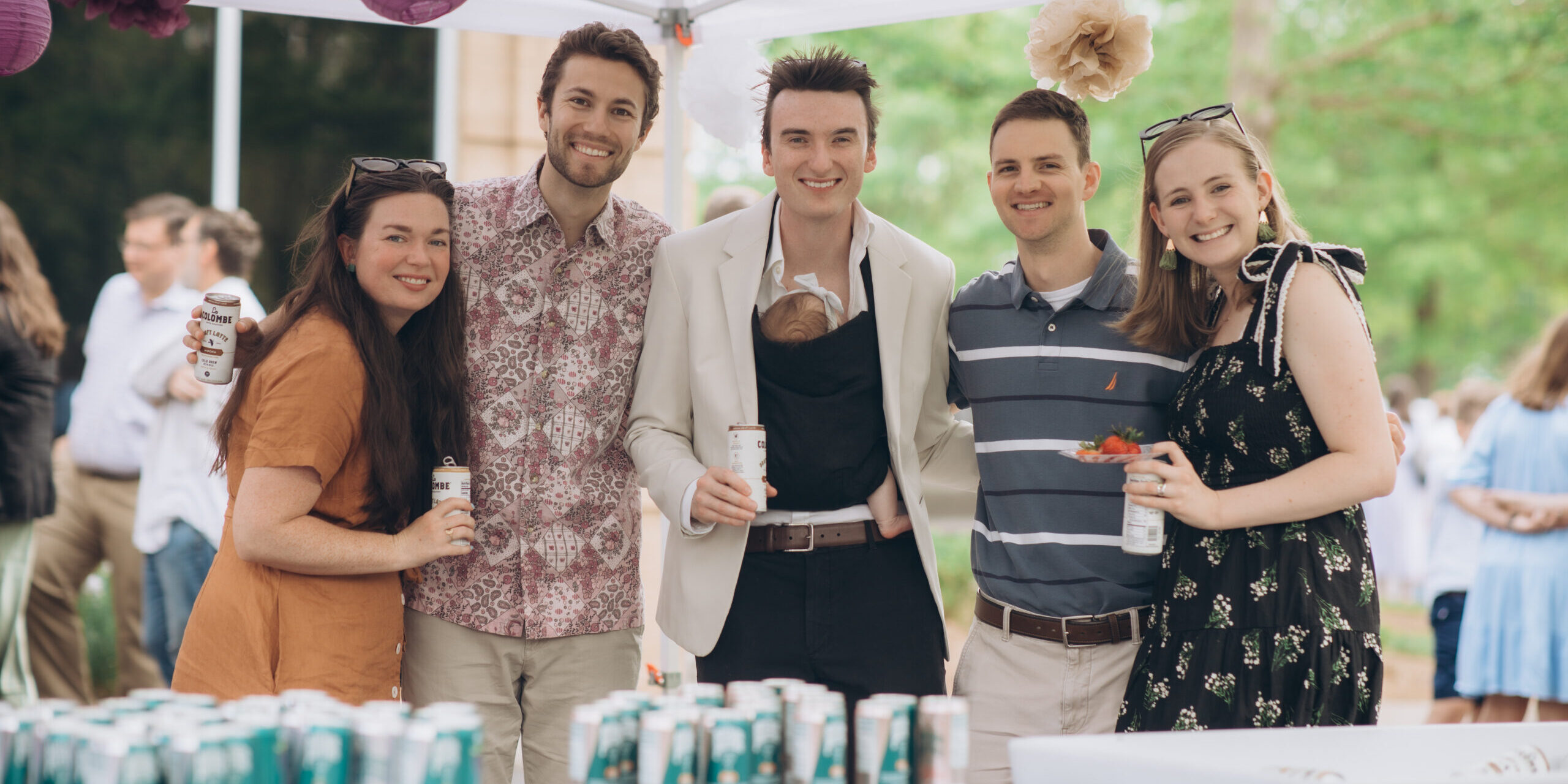 Five people stand together under a decorated tent at an outdoor event, holding drinks and smiling at the camera. One person is wearing a baby carrier with a baby inside. Cans are arranged on tables in front of them.