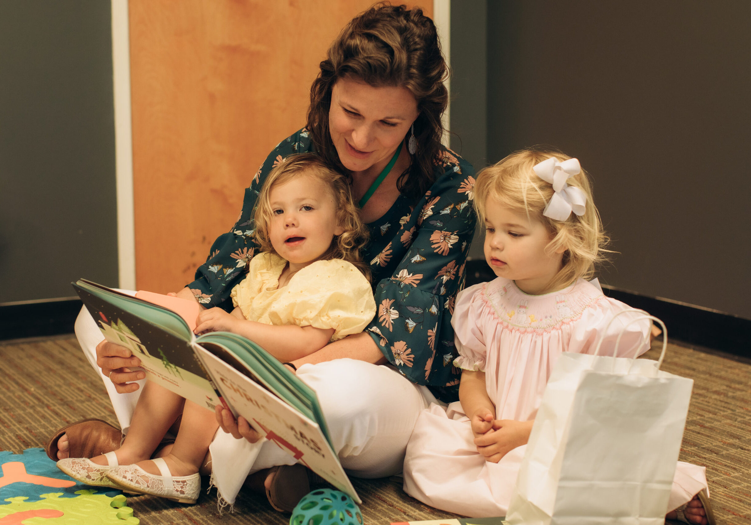 A woman sits on the floor reading a book to two young girls. One girl sits on her lap, and the other sits beside her holding a white bag. Colorful toys and a mat are nearby. All three are focused on the book.