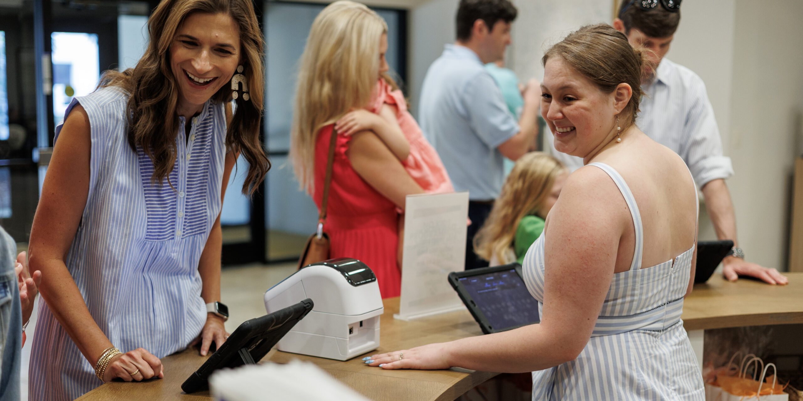 Two women in light summer dresses smile and interact at a reception desk with tablets and a printer. Other people, including a child, are gathered in the background inside a bright, modern space.