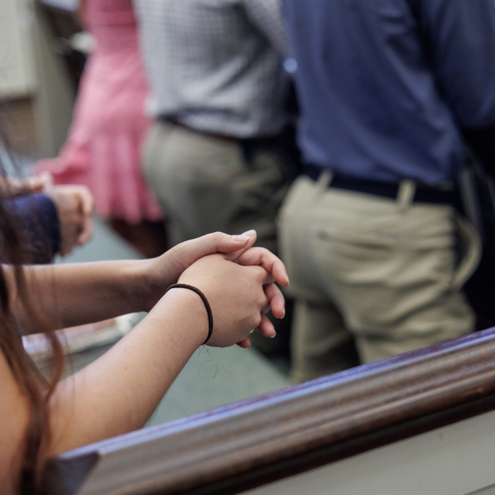 A person with long hair clasps their hands together in front of them, standing at a wooden pew among other people, some dressed in formal or semi-formal attire, in what appears to be a church or similar setting.