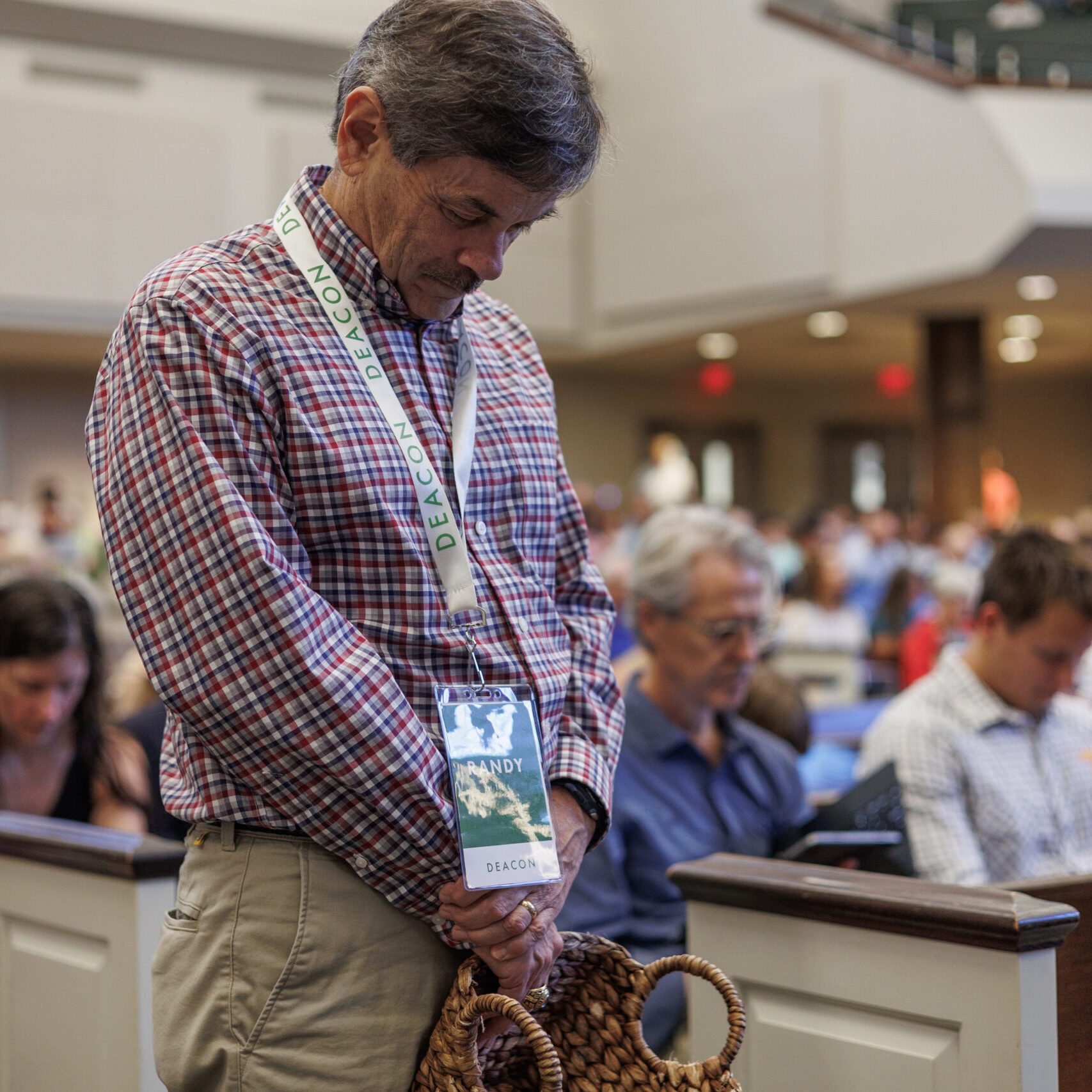 A man with a conference badge stands in a crowded room, bowing his head in prayer. Others around him are also seated with heads bowed, suggesting a solemn moment during an event or gathering.