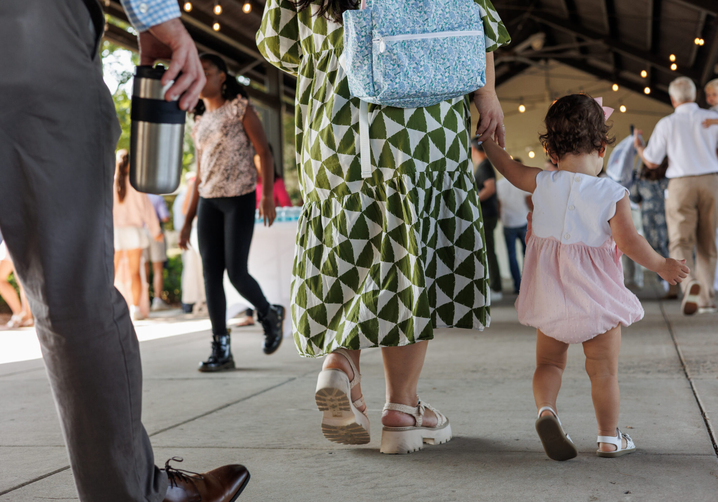 A toddler holding hands with an adult wearing a green patterned dress walks under a pavilion with string lights, surrounded by people at a busy outdoor event.