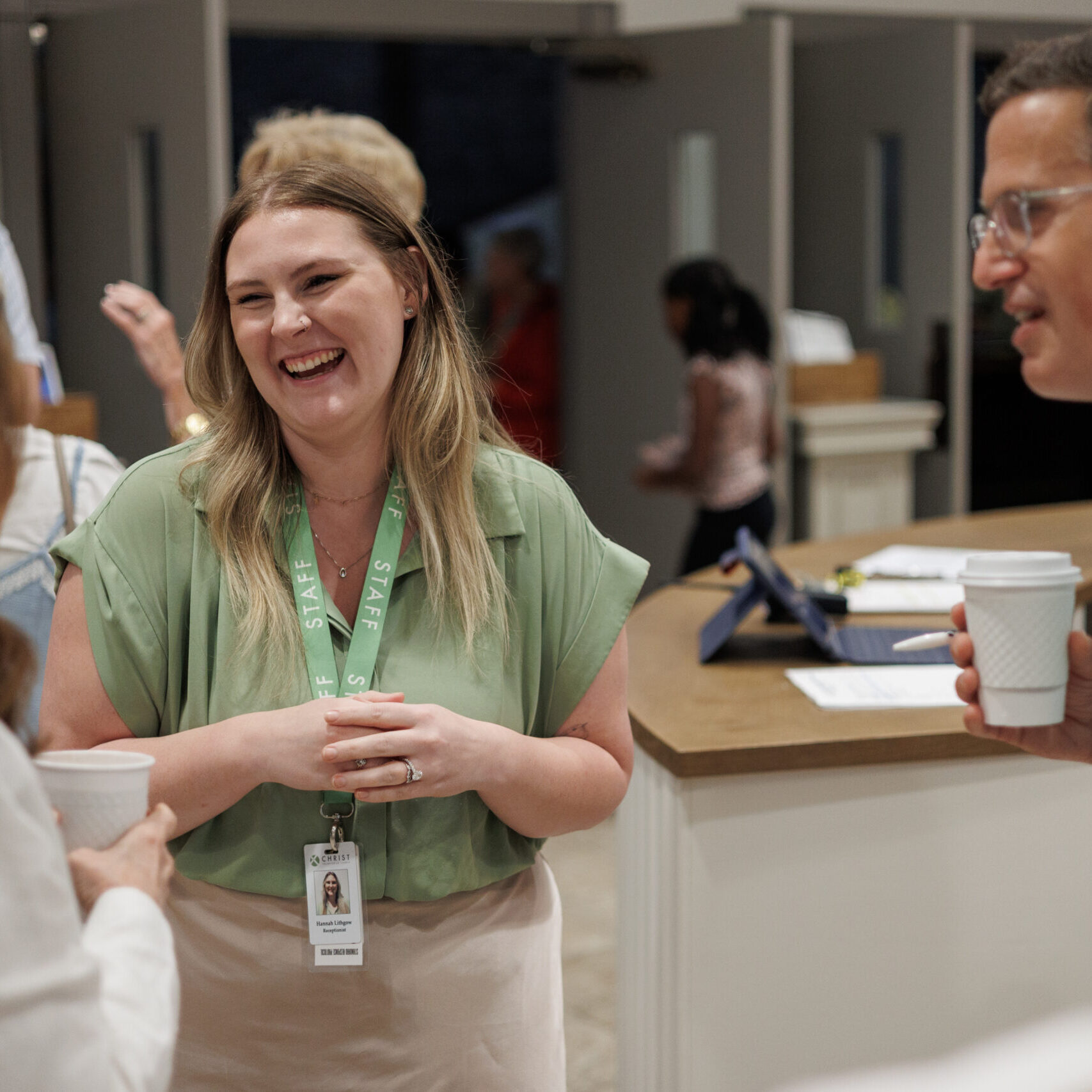 A woman wearing a green lanyard and name badge smiles and talks with two people holding coffee cups in a bright, indoor setting. Other people are visible in the background.