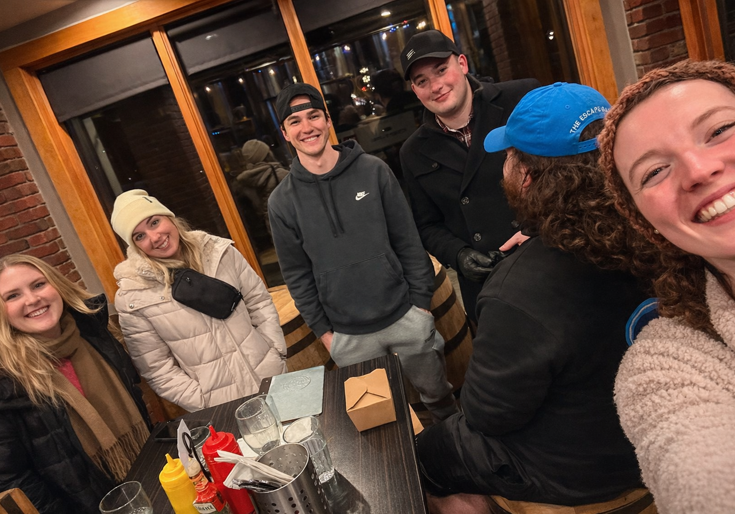 Six friends gather around a table at a cozy restaurant. Five are standing or sitting facing the camera, smiling, while one with curly hair and a blue cap has their back to the camera. Condiments and dishes are on the table.