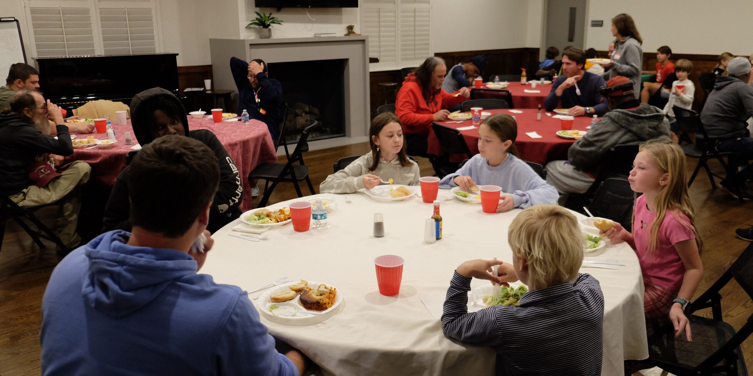 A group of children and adults sit around tables eating a meal together in a bright, indoor setting. Plates of food and red cups are on the tables, and people are engaged in conversation.