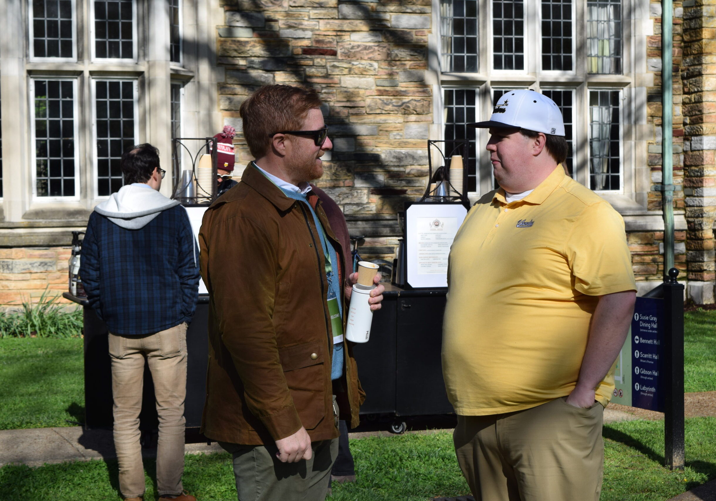 Two men stand talking outside on a sunny day; one wears a brown jacket and sunglasses, holding a cup, while the other wears a yellow polo shirt and white cap. Two other people and a stone building are seen in the background.