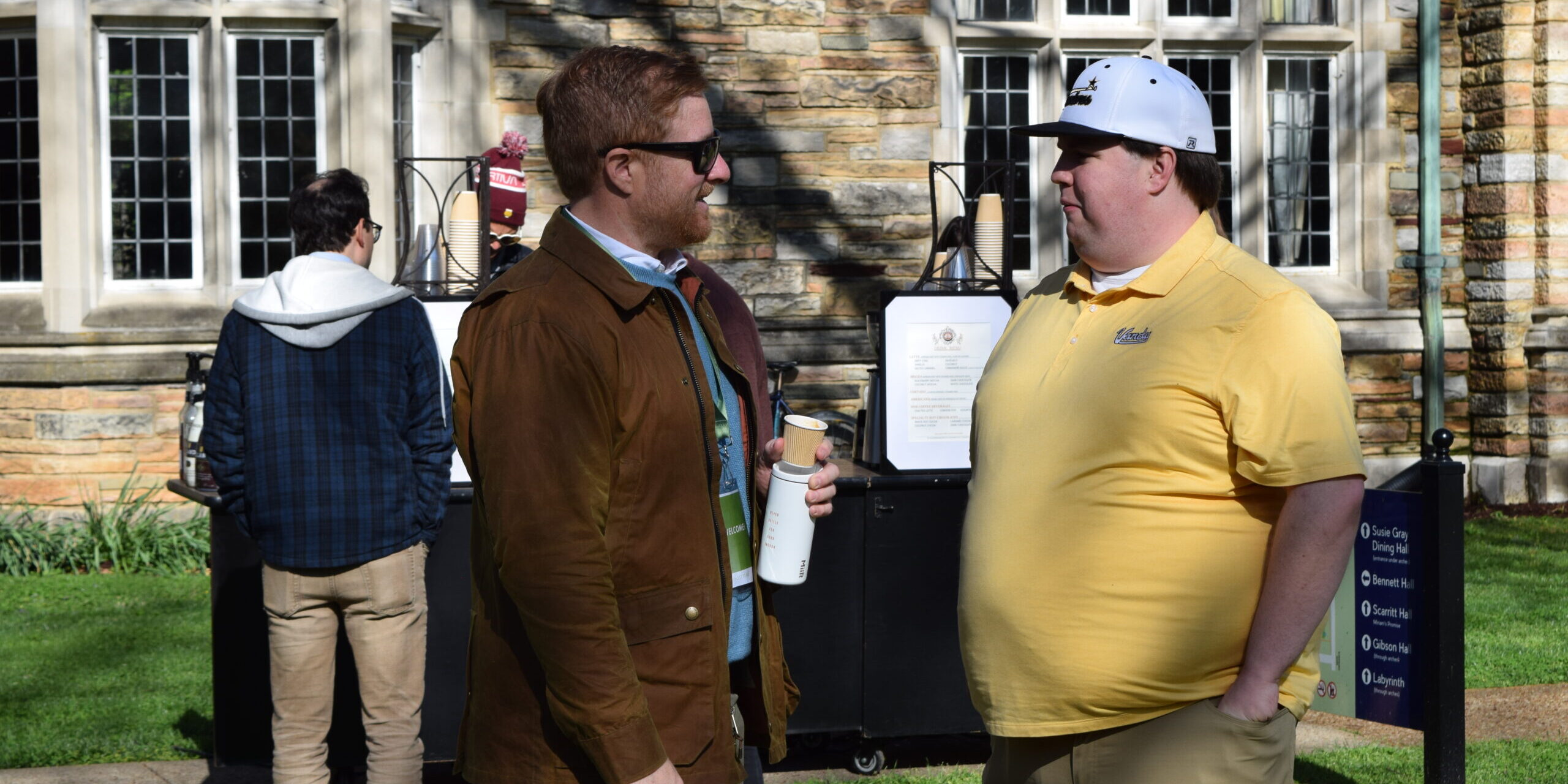 Two men stand talking outside on a sunny day; one wears a brown jacket and sunglasses, holding a cup, while the other wears a yellow polo shirt and white cap. Two other people and a stone building are seen in the background.