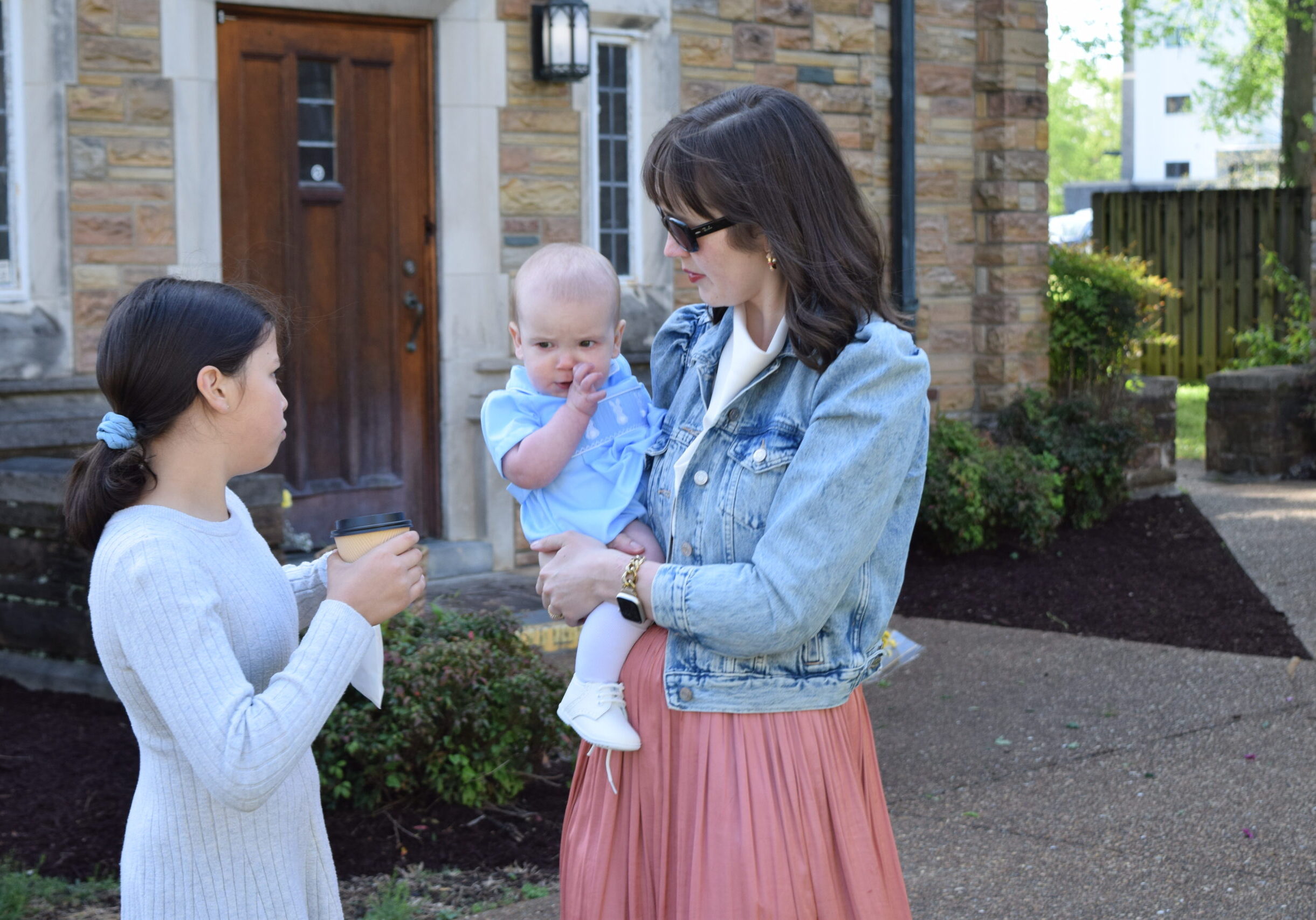 A woman in sunglasses holds a baby dressed in blue while talking to a young girl with a ponytail holding a cup, standing outside a stone building with a wooden door.