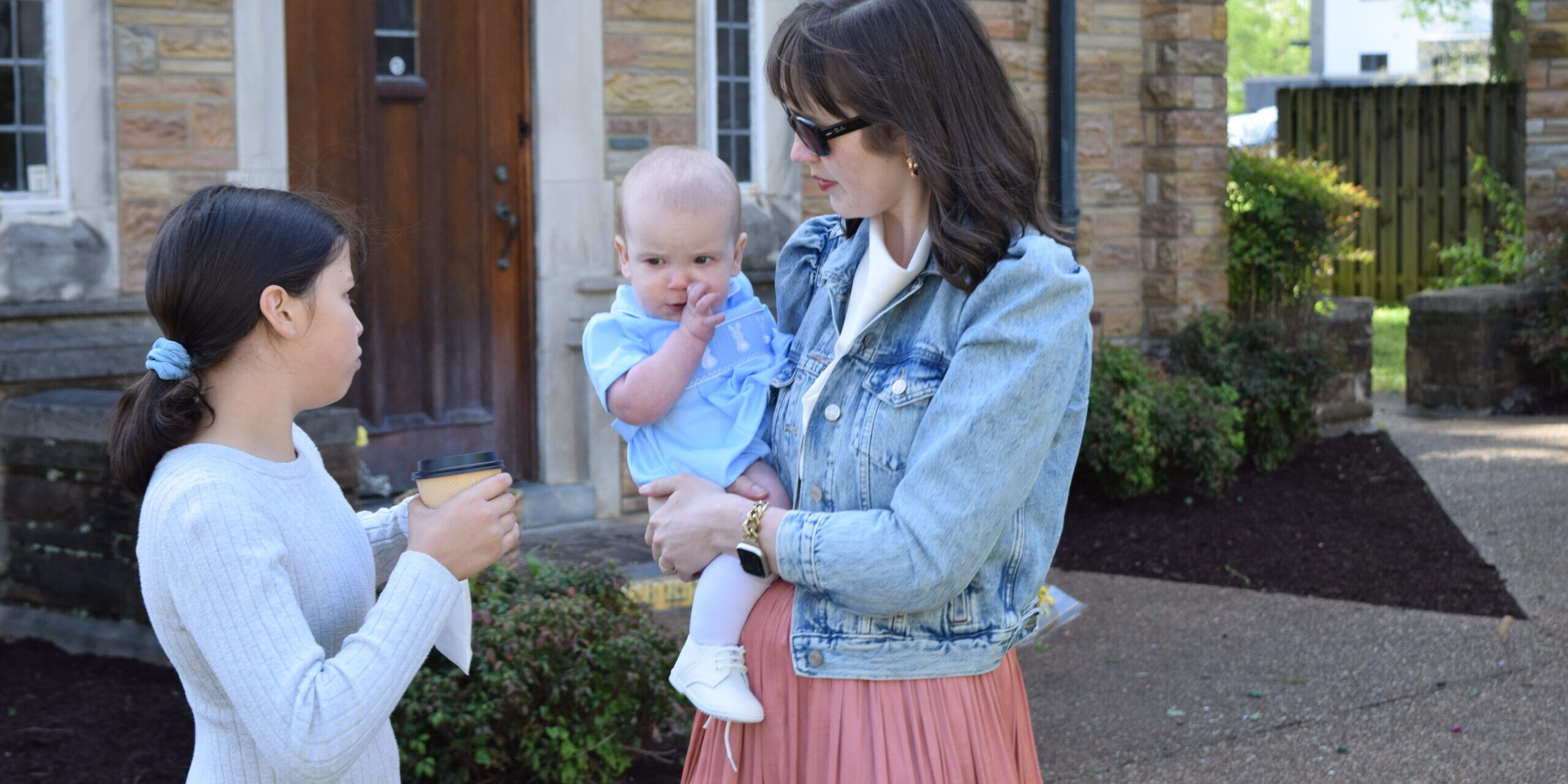 A woman in sunglasses holds a baby dressed in blue while talking to a young girl with a ponytail holding a cup, standing outside a stone building with a wooden door.