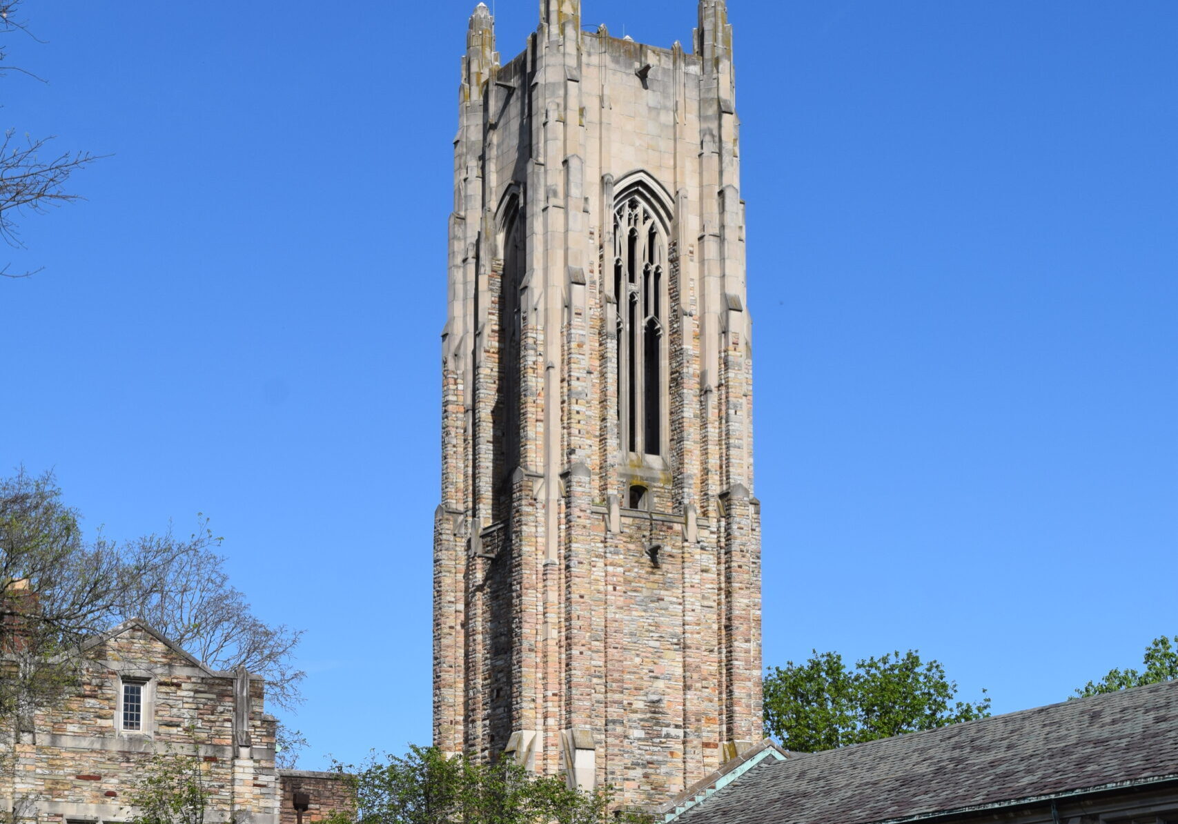 People stand and sit on a grassy lawn with picnic blankets and chairs in front of a tall stone church tower on a sunny day. Some are dressed formally, and the sky is clear and blue.