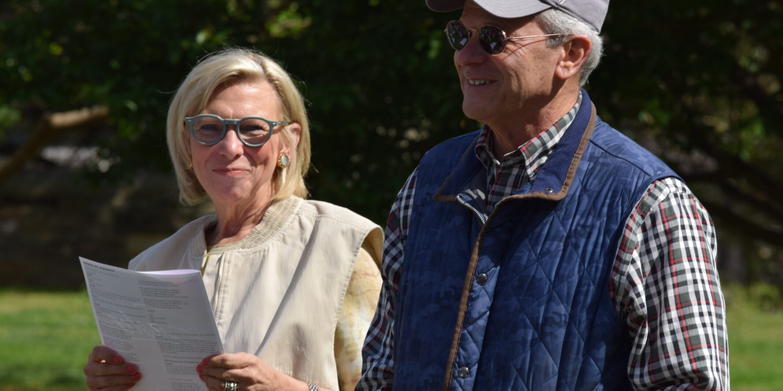 Two older adults stand outdoors. The woman, holding a paper, wears glasses and a light vest. The man wears sunglasses, a gray cap, a blue quilted vest, and a plaid shirt. Both are smiling in bright daylight.
