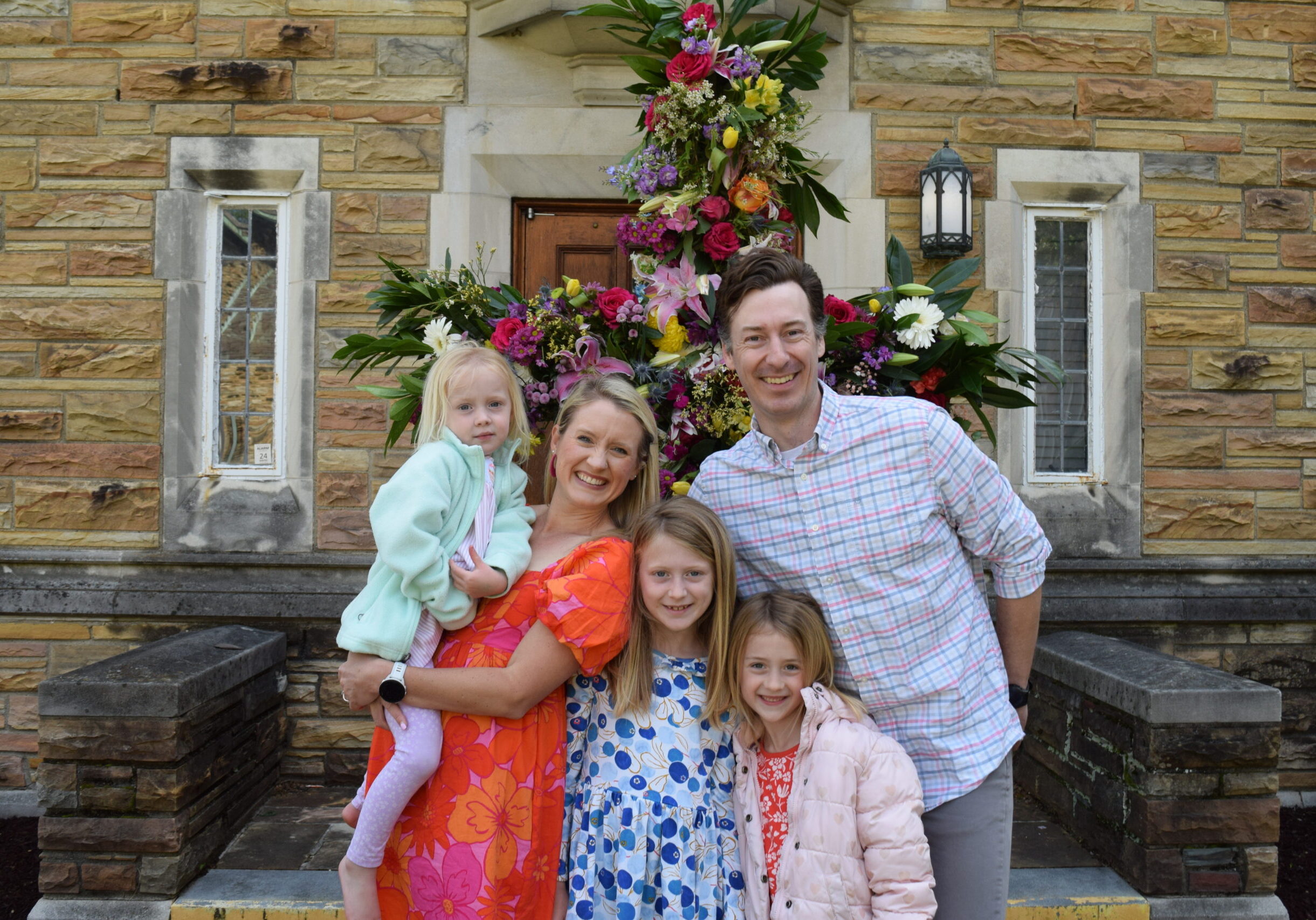 A smiling family of five poses in front of a stone building with large windows and a colorful floral arrangement. The parents hold their three daughters, who are wearing bright, patterned dresses and jackets.