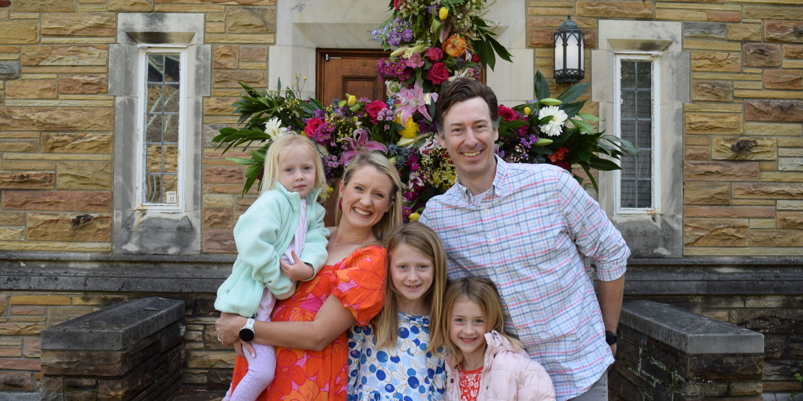 A smiling family of five poses in front of a stone building with large windows and a colorful floral arrangement. The parents hold their three daughters, who are wearing bright, patterned dresses and jackets.