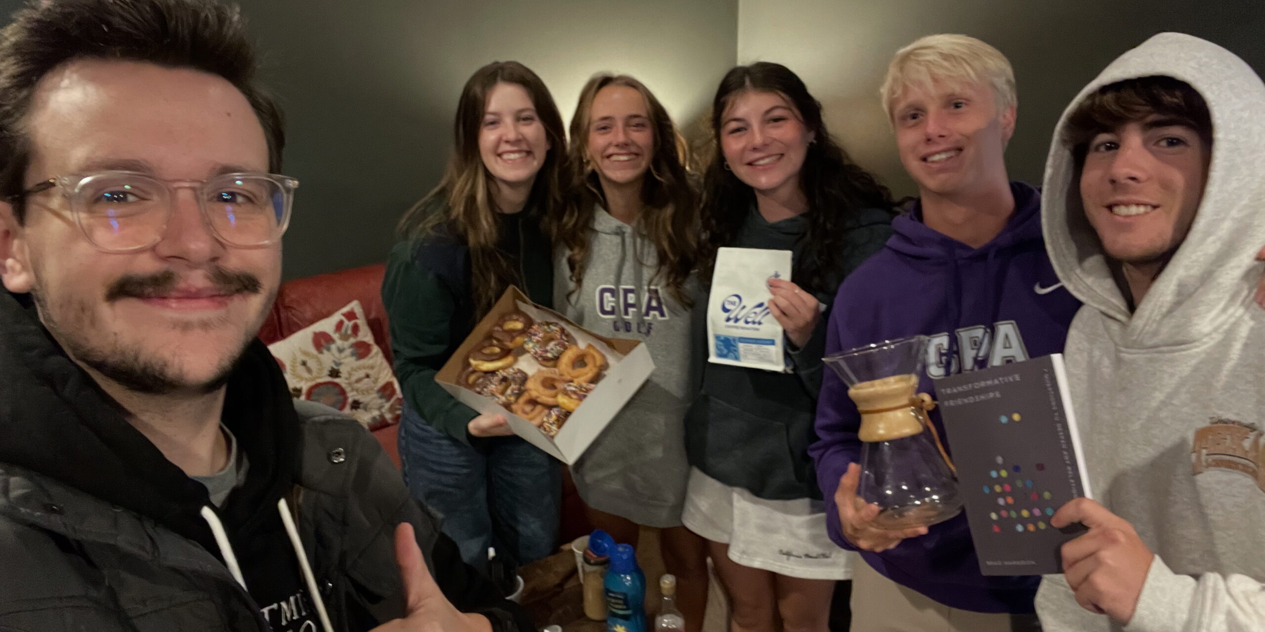 A group of five smiling young adults pose indoors. One person holds a box of donuts, another holds a Chemex coffee maker, and several hold books and papers. The group appears cheerful and relaxed.
