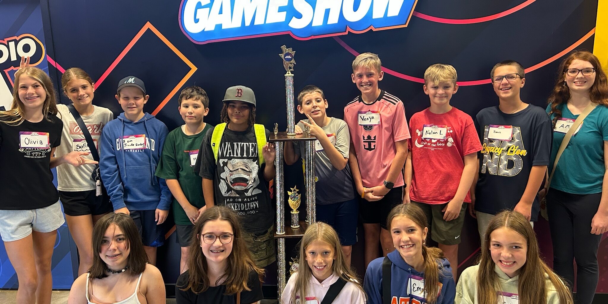 A group of smiling kids and teens pose indoors around a large trophy in front of a wall that reads Great Big Game Show. Most wear name tags and casual clothes.