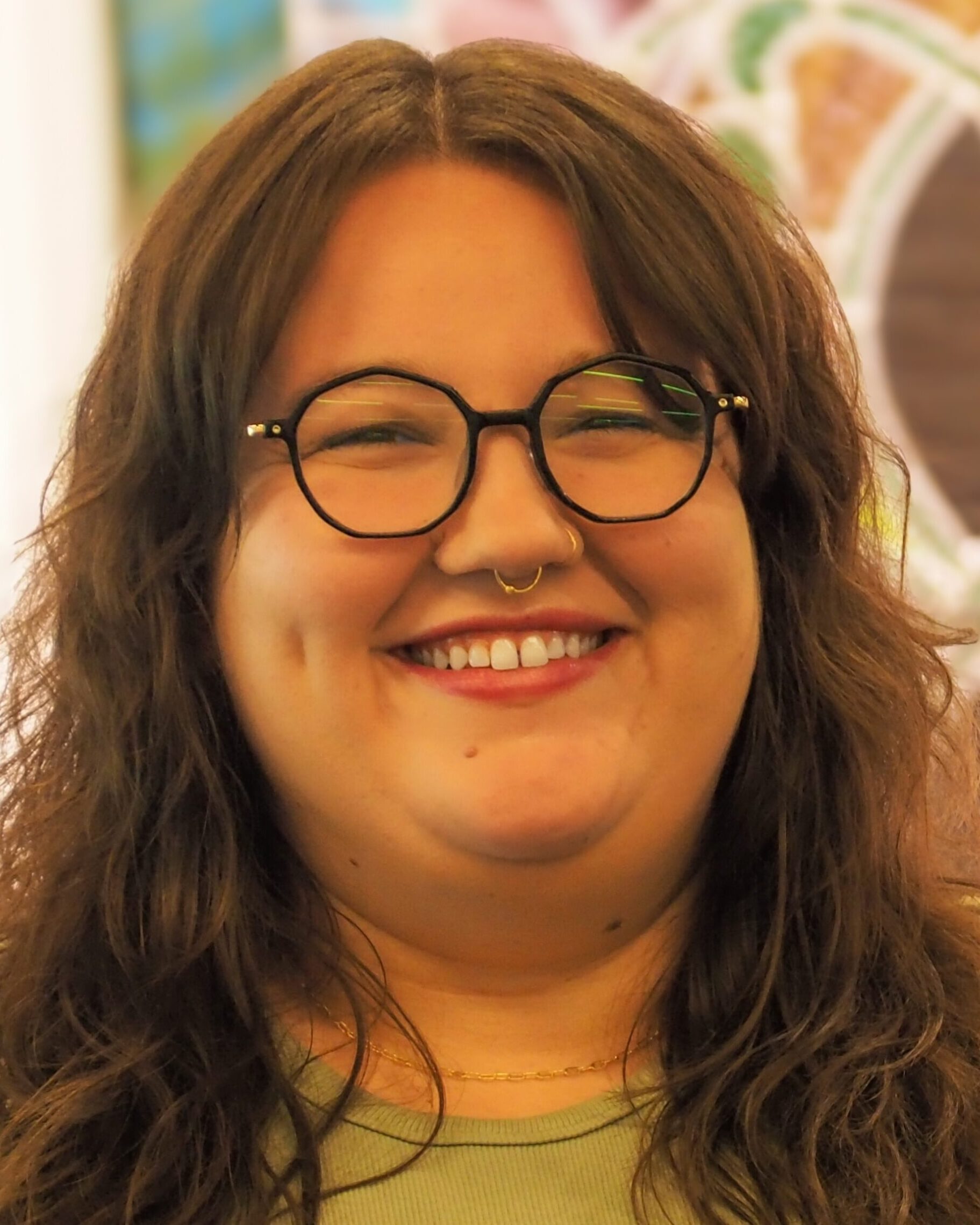 A person with long brown hair, glasses, and a septum ring smiles warmly at the camera. They are wearing a light green top and a gold necklace, with a blurred colorful background behind them.