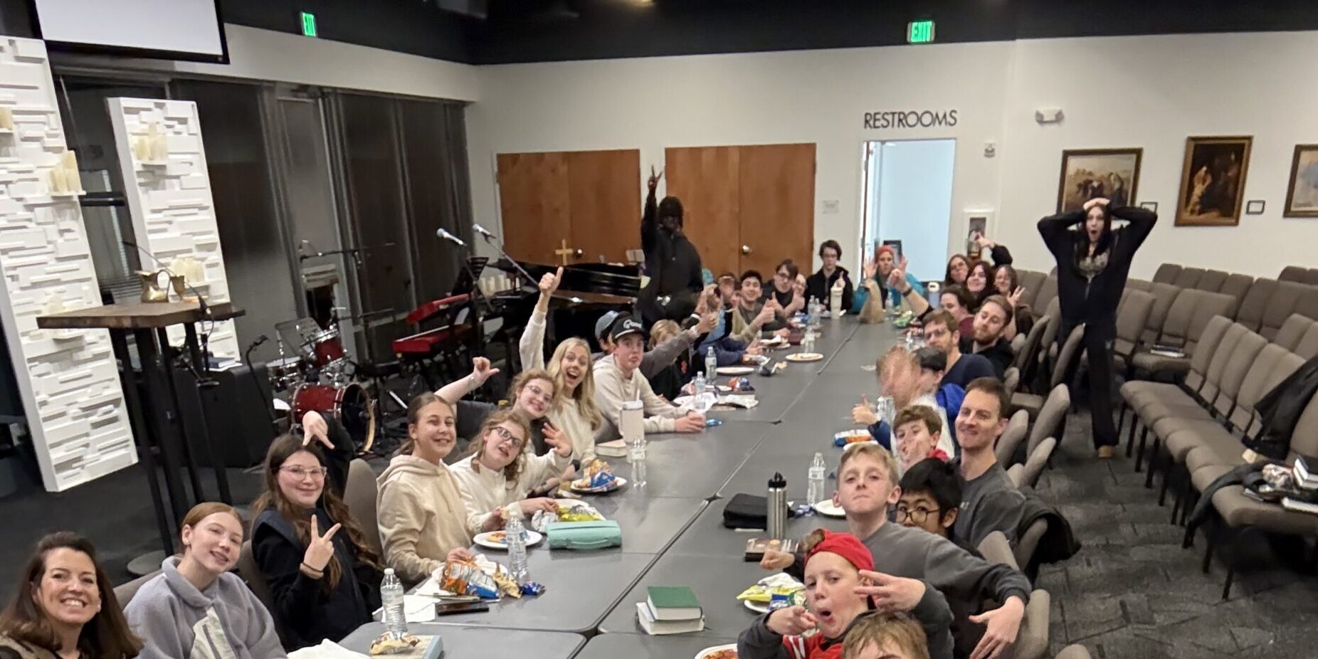 A large group of people, mostly young adults and teens, sit around long tables eating and smiling at the camera. A woman in front takes a selfie, and the room has rows of chairs and a Restrooms sign in the back.
