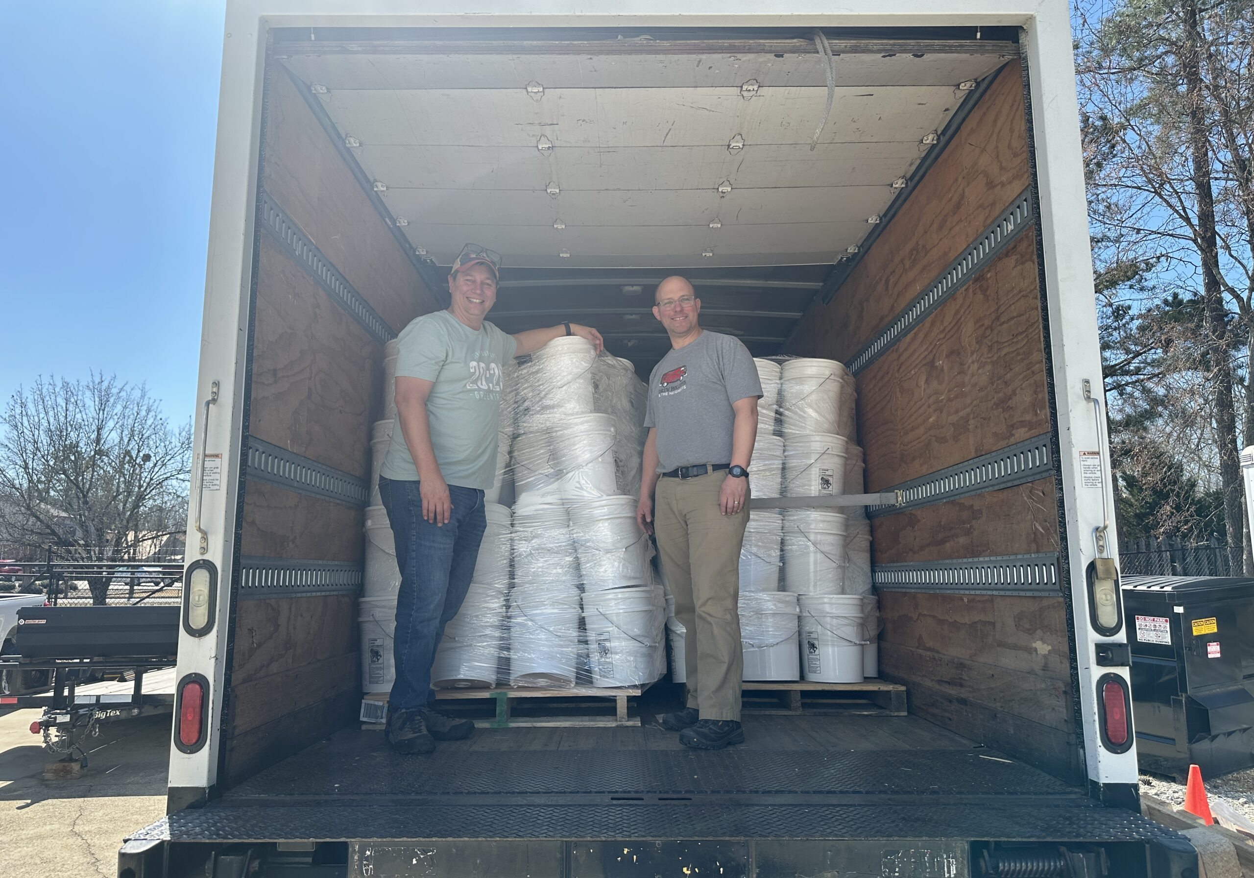 Two men stand inside the back of a box truck filled with large white bundles and buckets on pallets. The truck’s doors are open, and it is parked outdoors on a sunny day.