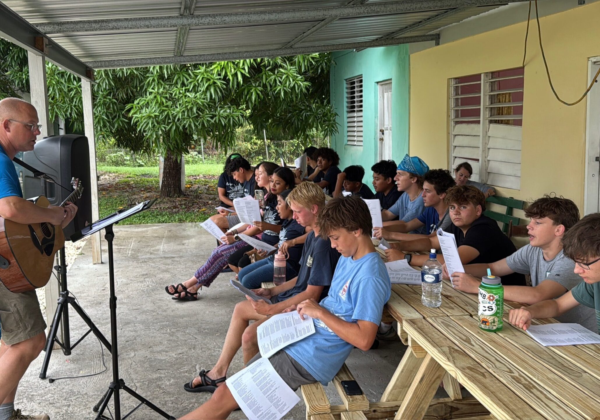 A man plays guitar and sings to a group of teenagers sitting outside under a covered patio. The students, holding song sheets, are seated on benches and picnic tables, listening and singing along. Trees and buildings are visible in the background.