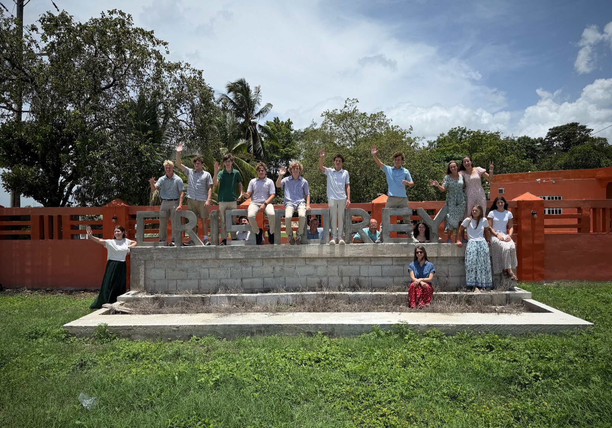A group of people stands and sits on steps in front of large block letters spelling FELIPE CARRILLO PUERTO with a bright orange fence behind them, waving and smiling on a sunny day.