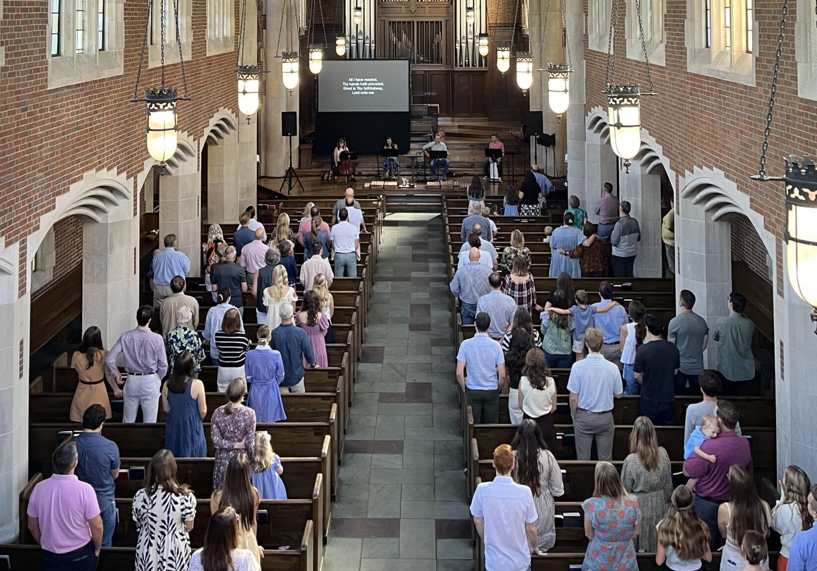A congregation stands in wooden pews of a large, brick-walled church with tall stained glass windows and a vaulted wooden ceiling. An organ is at the front above a screen displaying text.