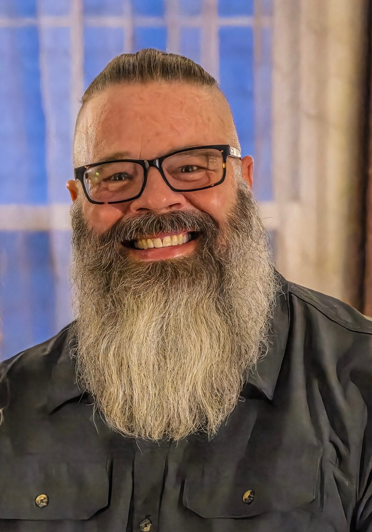 A smiling man with a large gray beard, glasses, and a dark shirt stands indoors in front of a window with curtains.