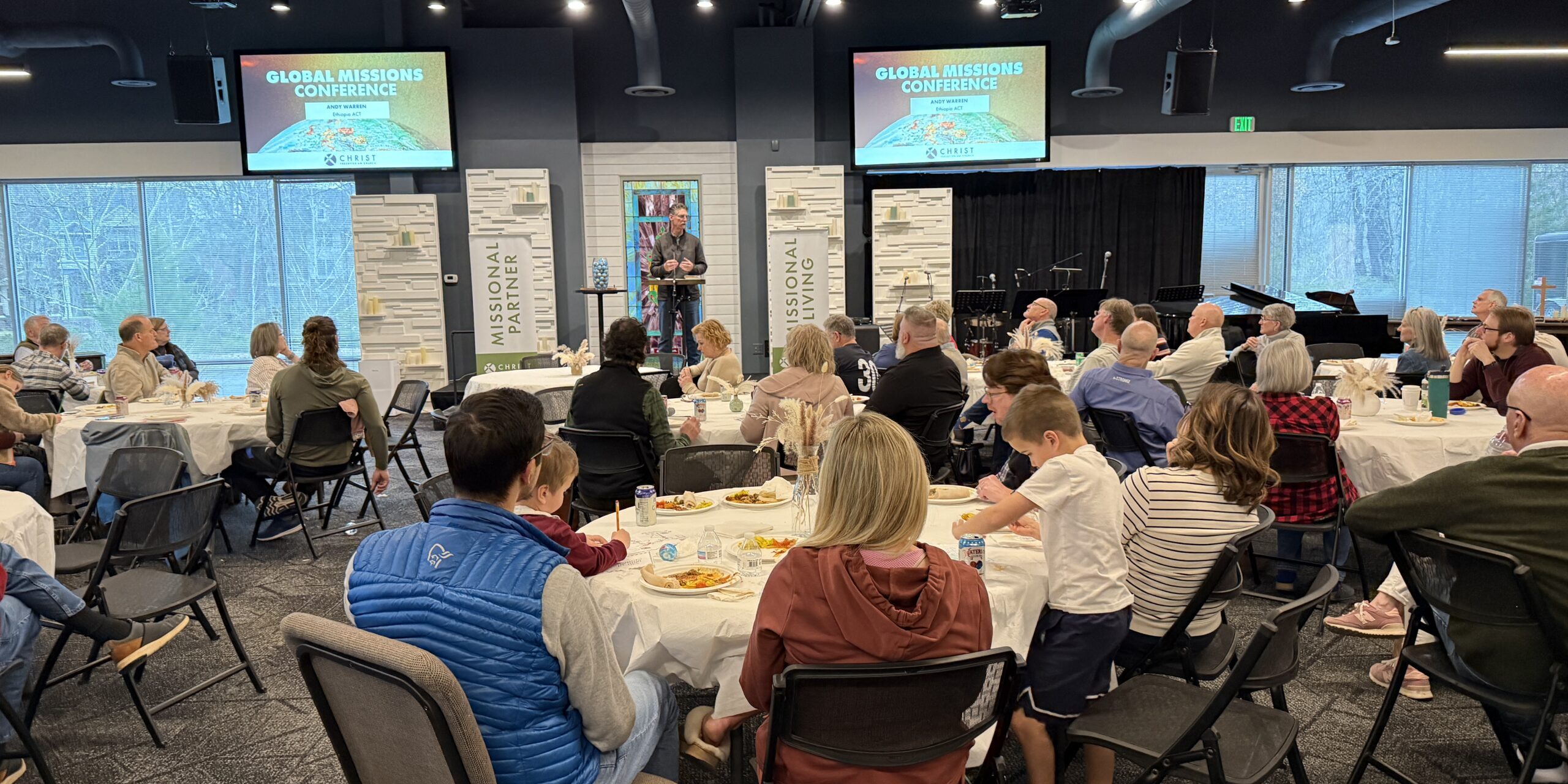 A group of people sit at round tables, eating and listening to a speaker at the front of a room during a Global Missions Conference. Two large screens display the event title. The setting appears modern with large windows.