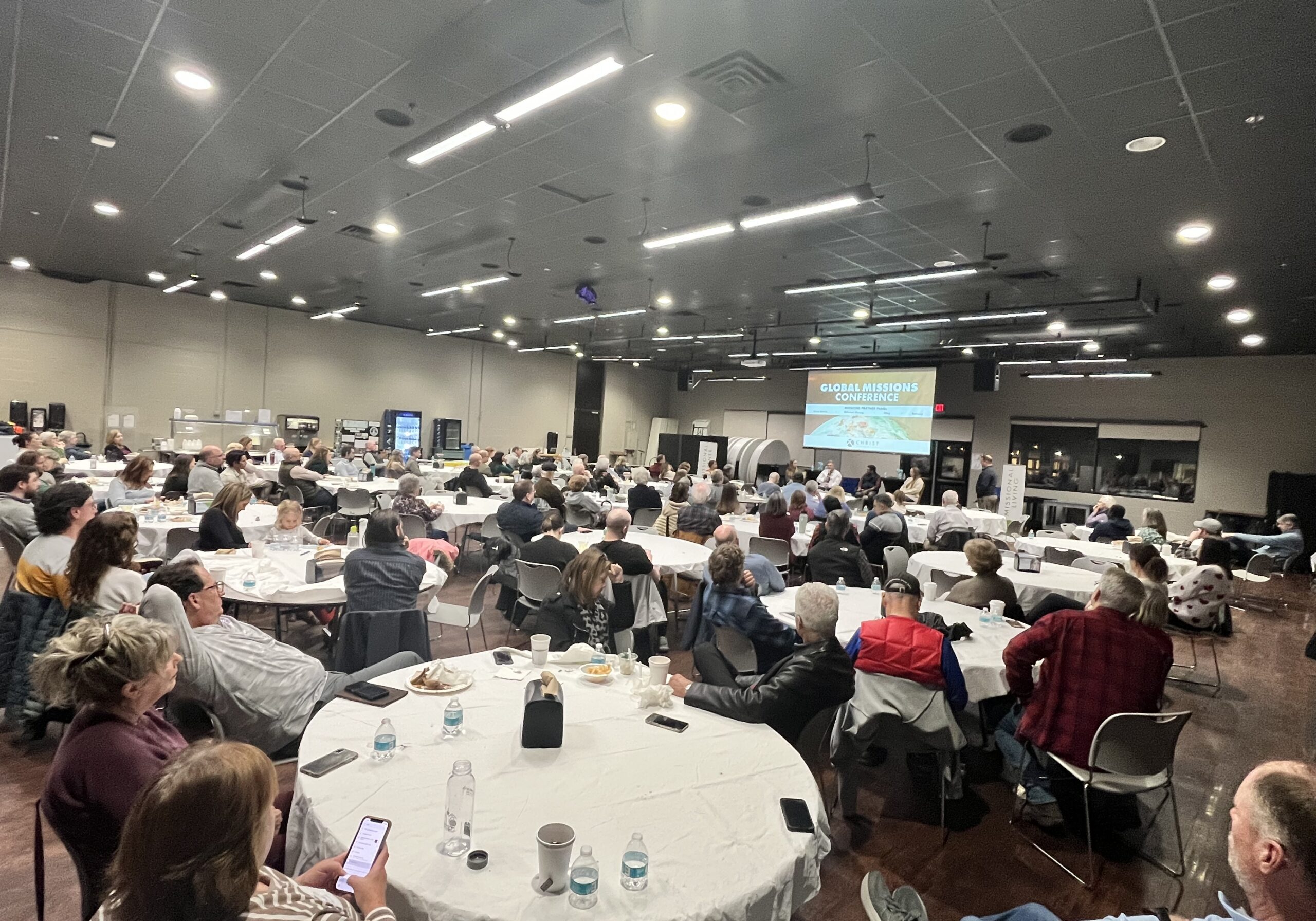 A large group of people sits at round tables in a conference room, facing a stage with a presentation screen displaying GLOBAL MISSIONS CONFERENCE. The room is well-lit and attendees are listening attentively.