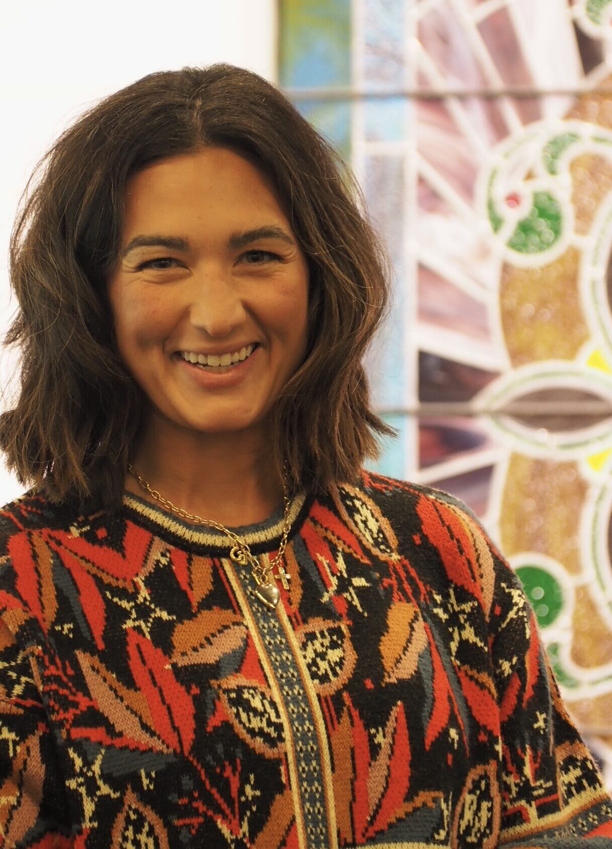 A woman with shoulder-length dark hair and a colorful patterned sweater smiles in front of a stained glass window with abstract designs.