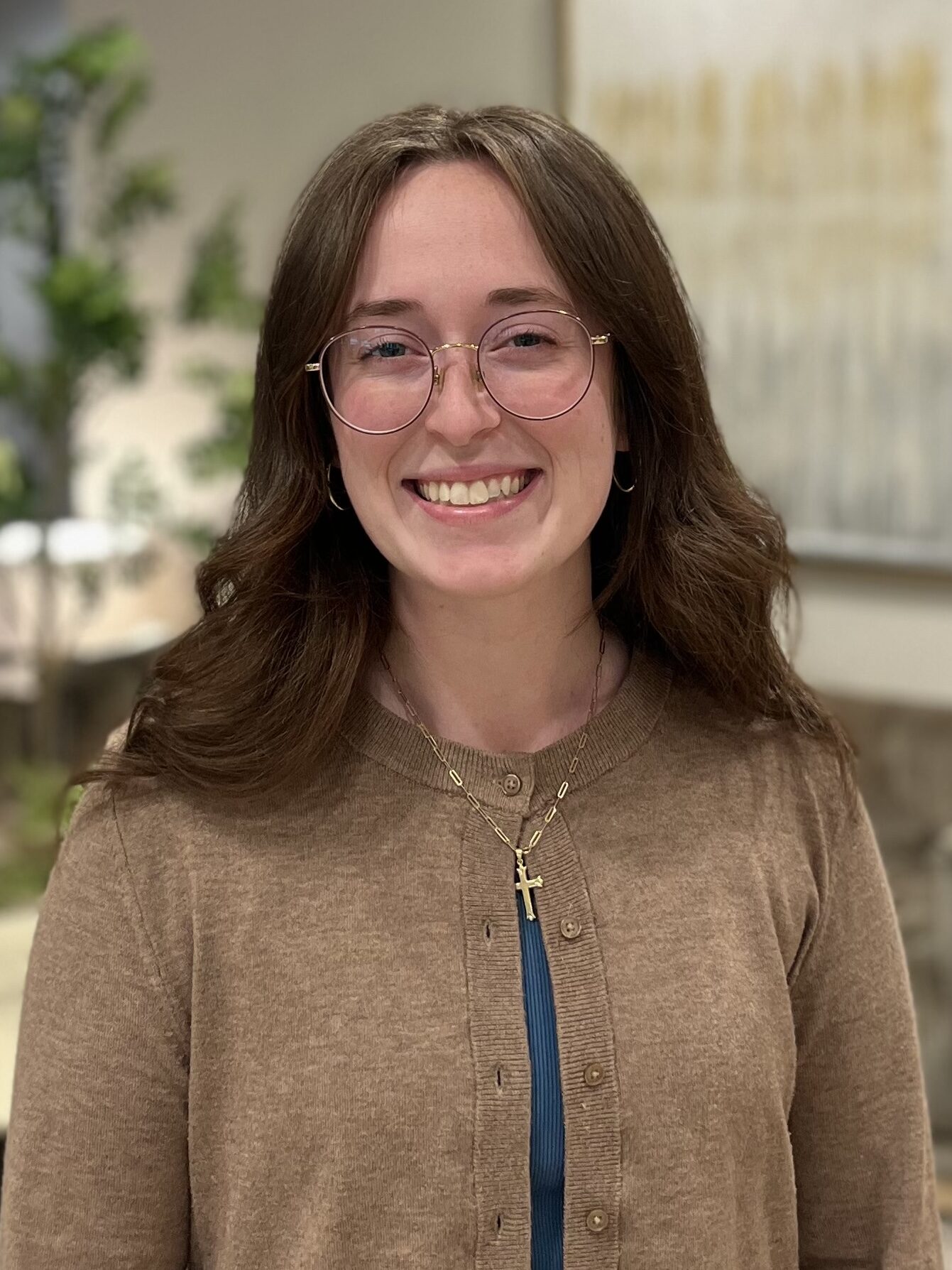 A young woman with long brown hair and round glasses smiles at the camera. She is wearing a tan cardigan over a blue top and a gold necklace with a cross pendant. A blurred indoor setting is in the background.