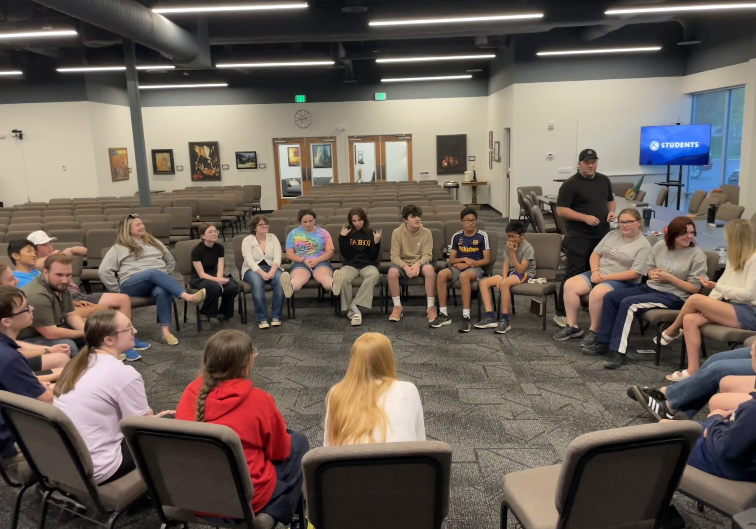 A group of teenagers and adults sit in a large circle of chairs inside a spacious room, engaged in conversation or activity, with a TV screen in the background displaying the word “STUDENTS.”.