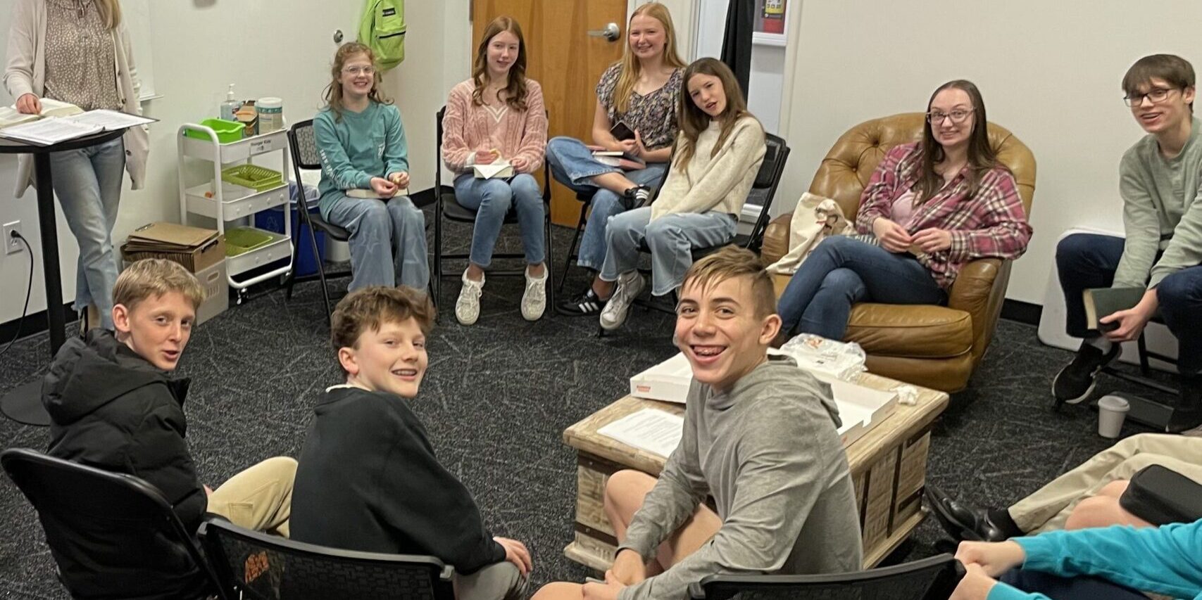 A group of teens sit in a circle in a cozy, well-lit classroom, some on couches and others on chairs. A woman stands by a whiteboard, smiling, while string lights hang over the group. Everyone looks engaged and happy.