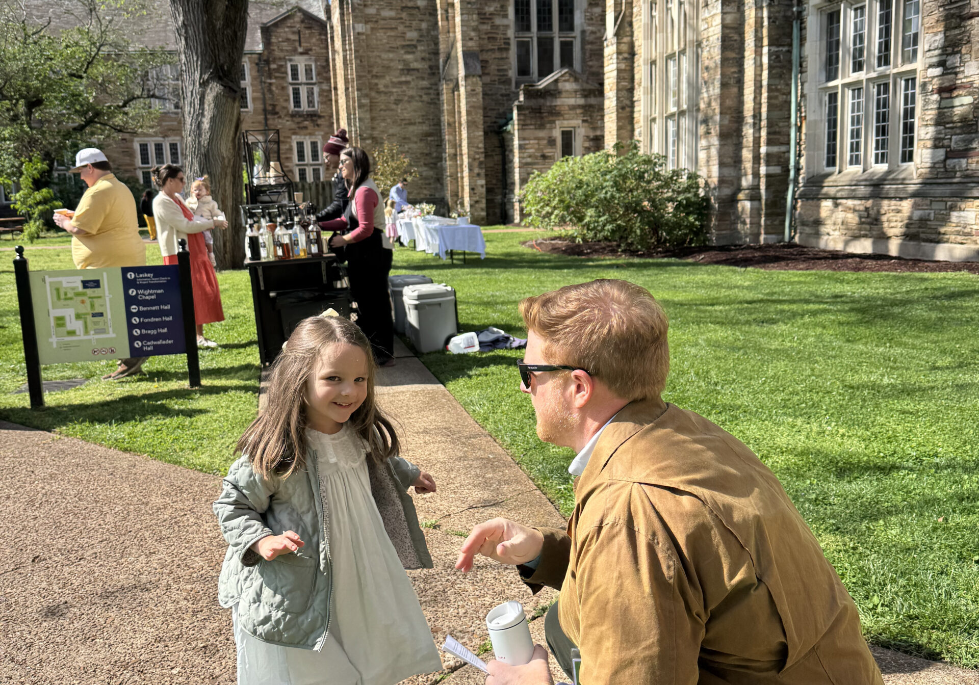 A man kneels to talk to a smiling young girl in a light dress on a sunny sidewalk outside a large stone building, with people gathered near a refreshment table in the background.