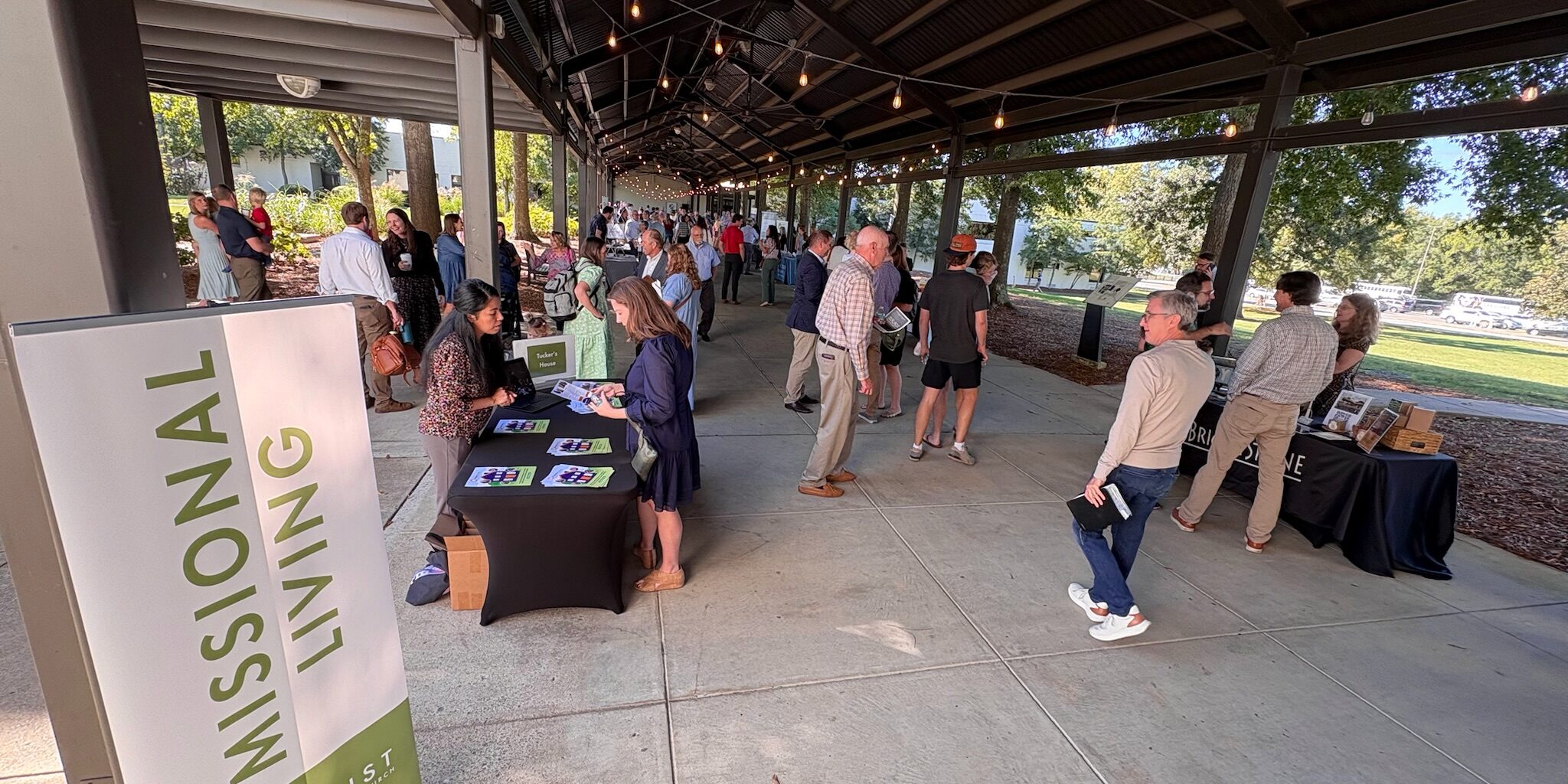 People gather under a covered outdoor area with informational tables set up. A vertical sign reads Missional Living. Sunlight filters through trees in the background. Some people are engaged in conversation or looking at displays.