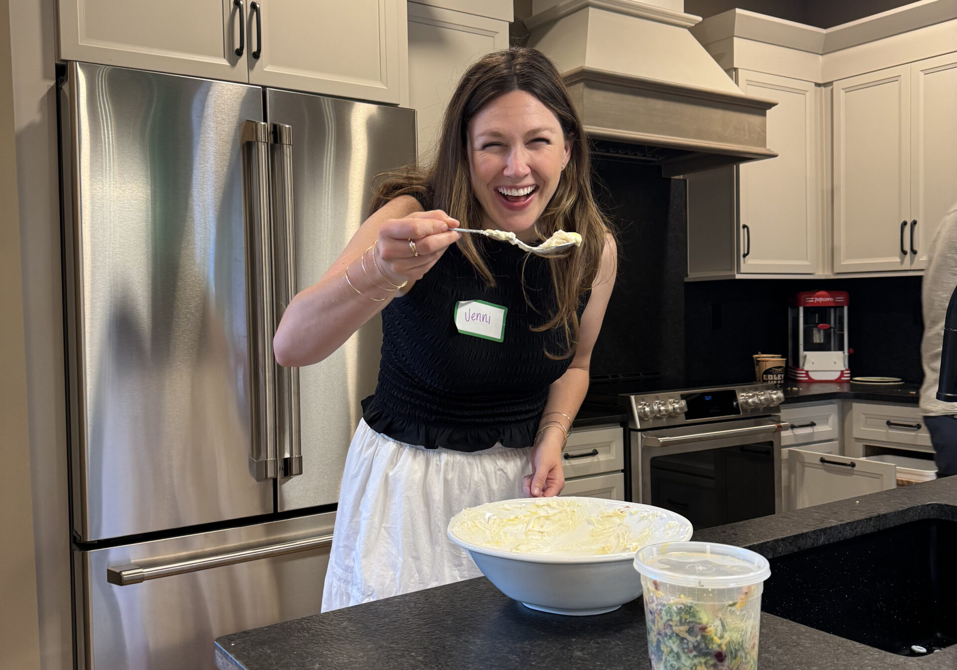 A woman smiling in a kitchen, holding a spoon with food above a large mixing bowl. There is a can of soda, a plastic container of salad, and tape on the black countertop in front of her.