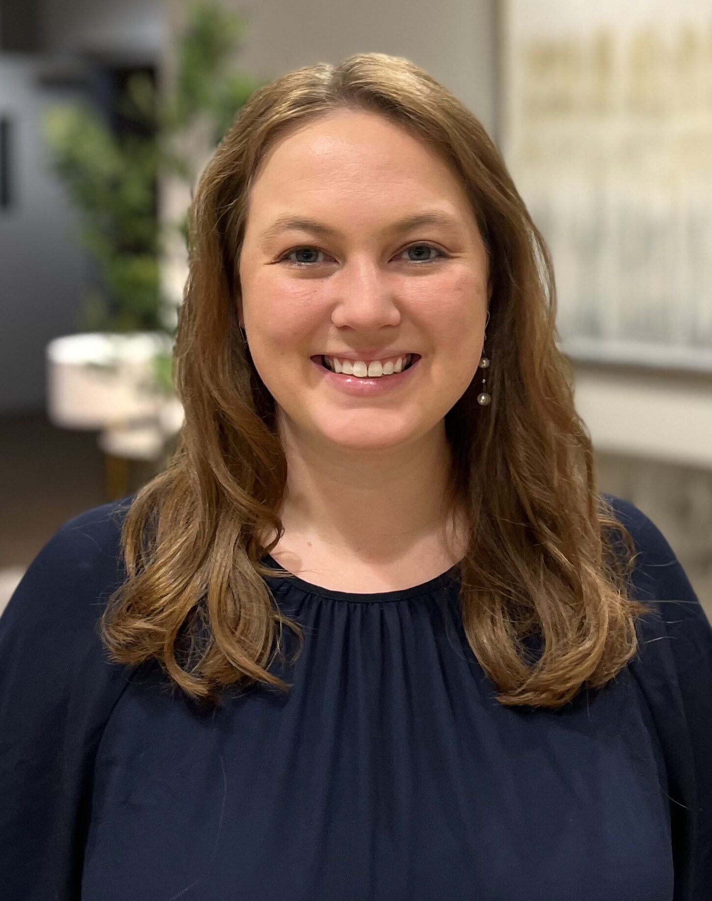 A woman with long light brown hair wearing a navy blue top is smiling at the camera. The background is softly blurred, showing indoor decor with plants and framed artwork.