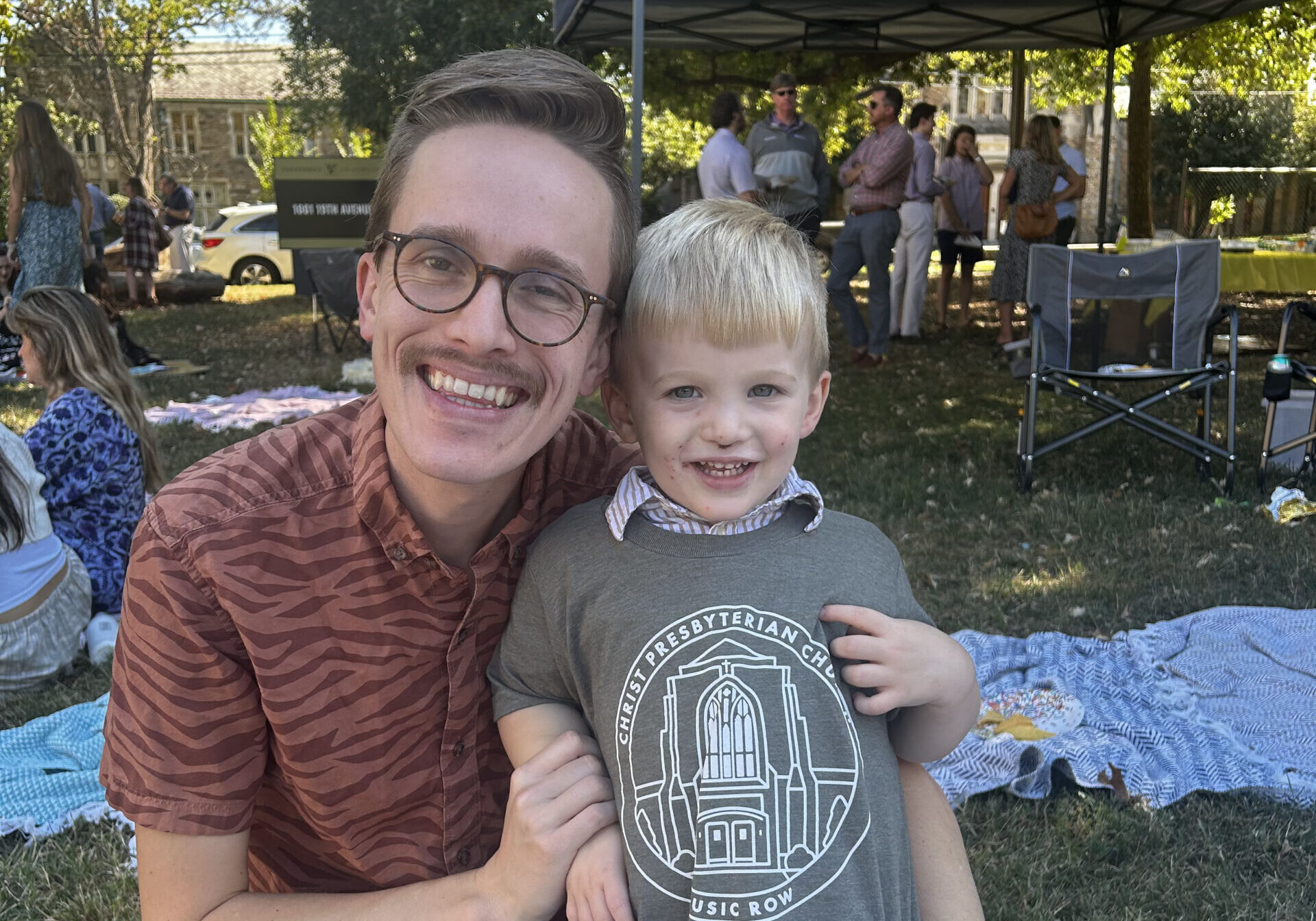 A smiling adult with glasses kneels next to a blonde boy outdoors on a sunny day. They are surrounded by picnic blankets and people, with a blue canopy tent in the background. The boy holds up his oversized t-shirt, smiling.