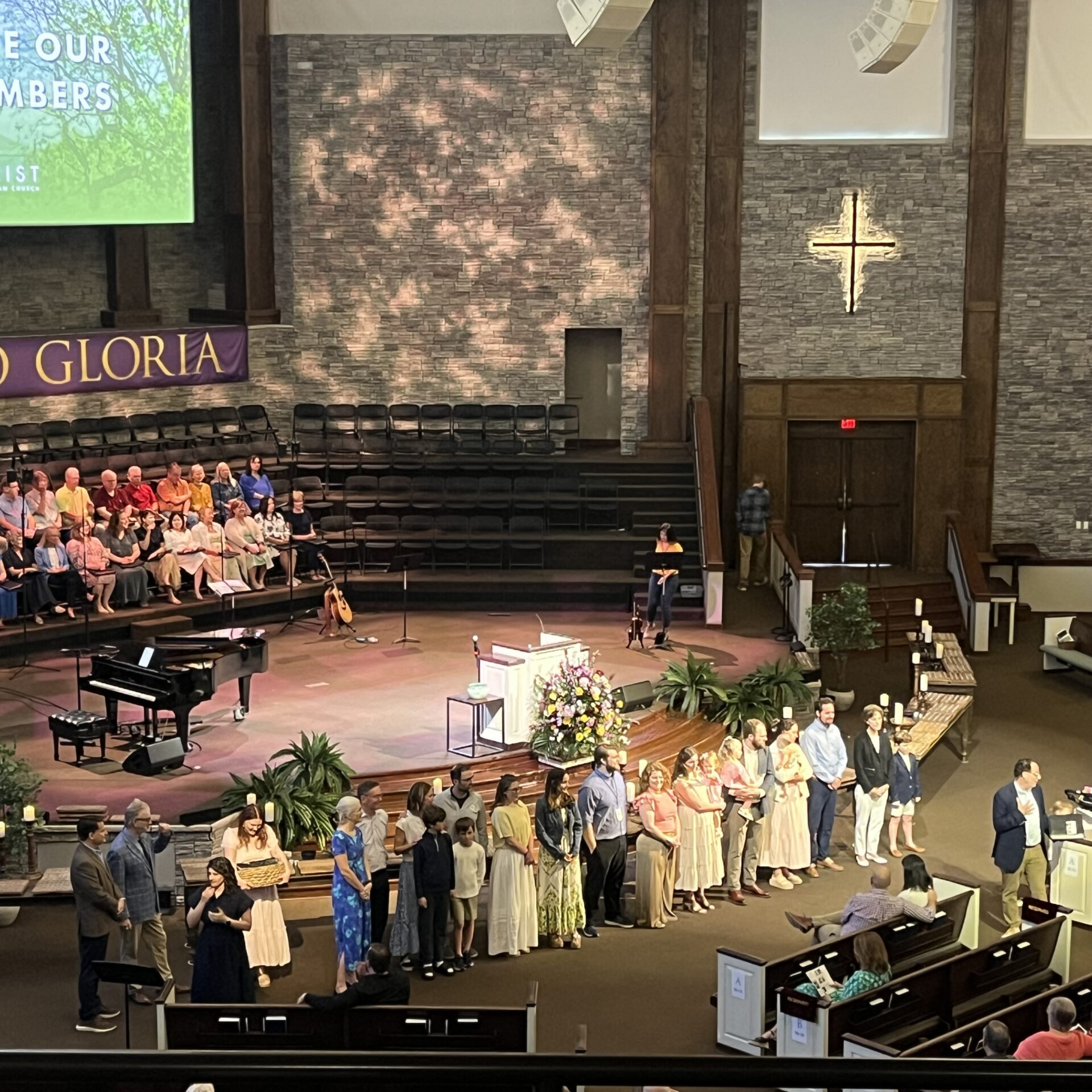 A church sanctuary with a group of people standing in front of the congregation. A screen reads Welcome Our New Members. The choir is seated on the left, and attendees are seated in pews. Flowers and instruments are near the stage.