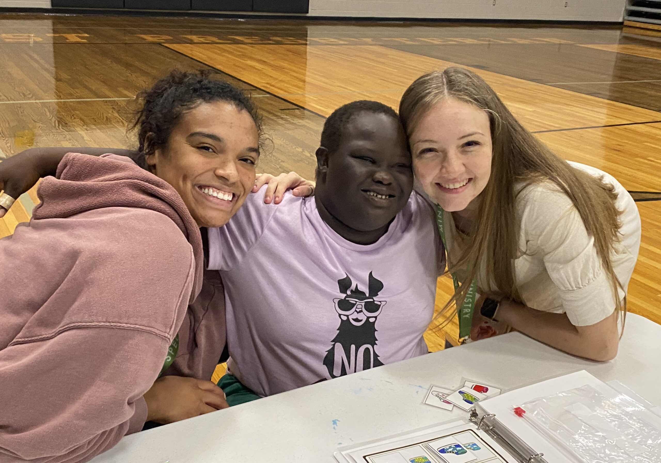Three smiling people sit closely together at a table in a gym, arms around each other. The person in the middle wears a light purple shirt with a llama graphic, and the others wear casual clothes. Papers and cards are on the table.