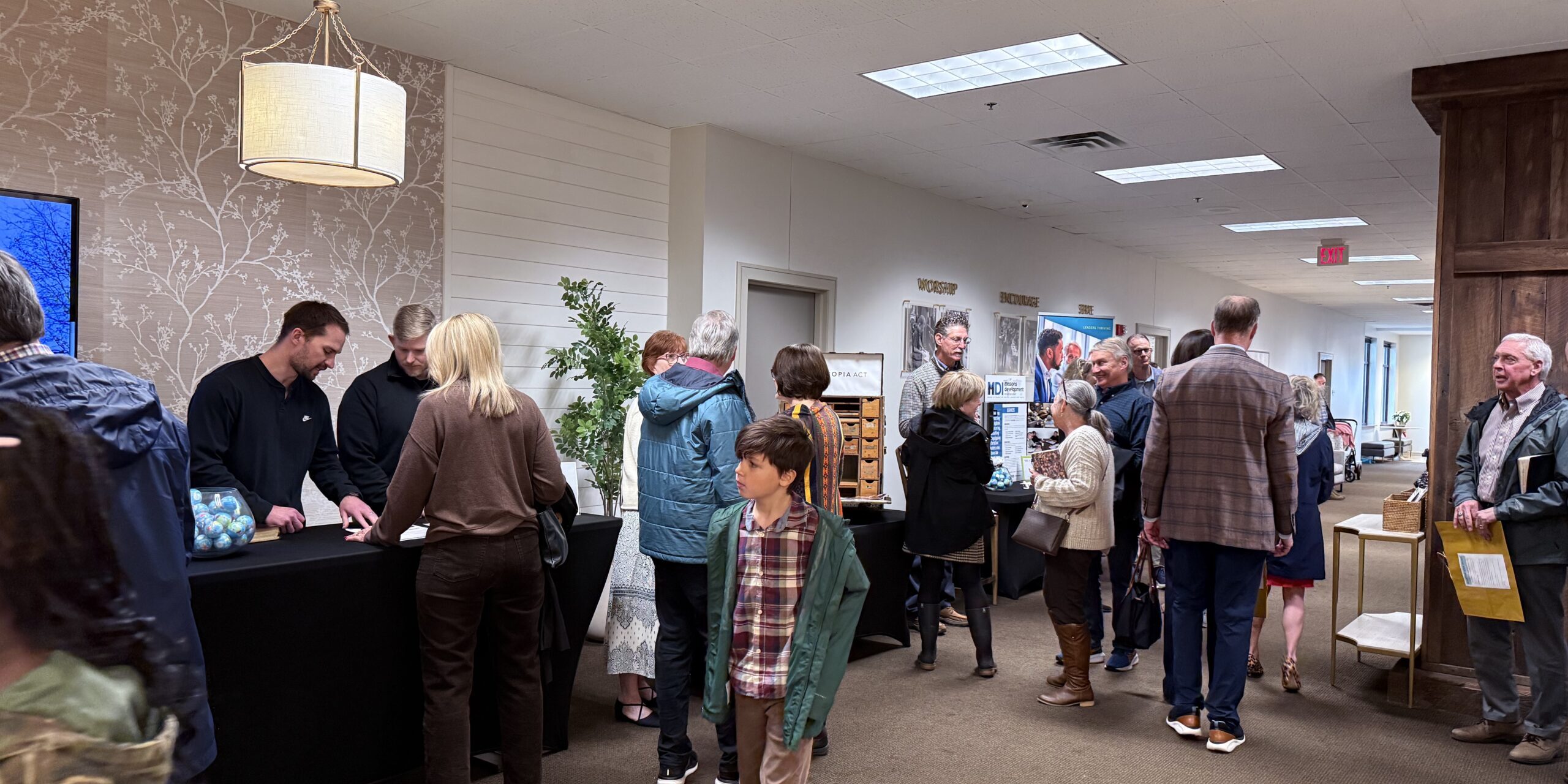 People gather and converse in a hallway at an indoor event. Some stand in line at a table with items, while others walk or chat in groups. The setting appears casual, with adults and children present.