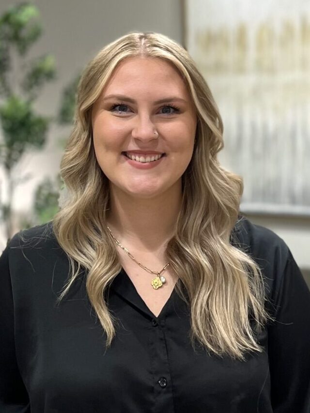A woman with long blonde hair wearing a black blouse and gold necklace smiles at the camera. The background is softly blurred with greenery and a light-colored painting.