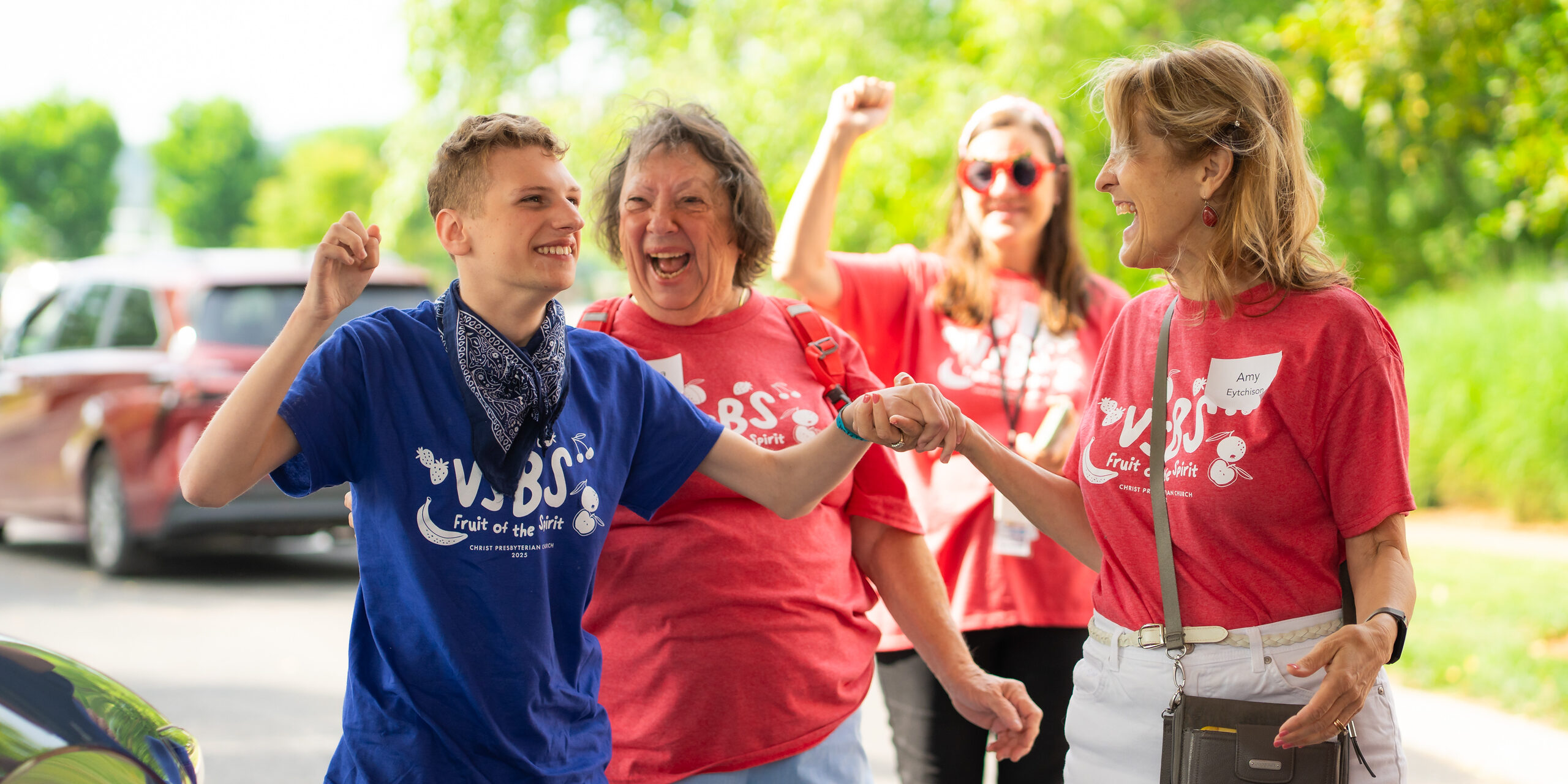 A young boy and three women, all smiling and wearing colorful VBS T-shirts, stand outdoors in the sunlight, celebrating and holding hands. Green trees and parked cars are visible in the background.