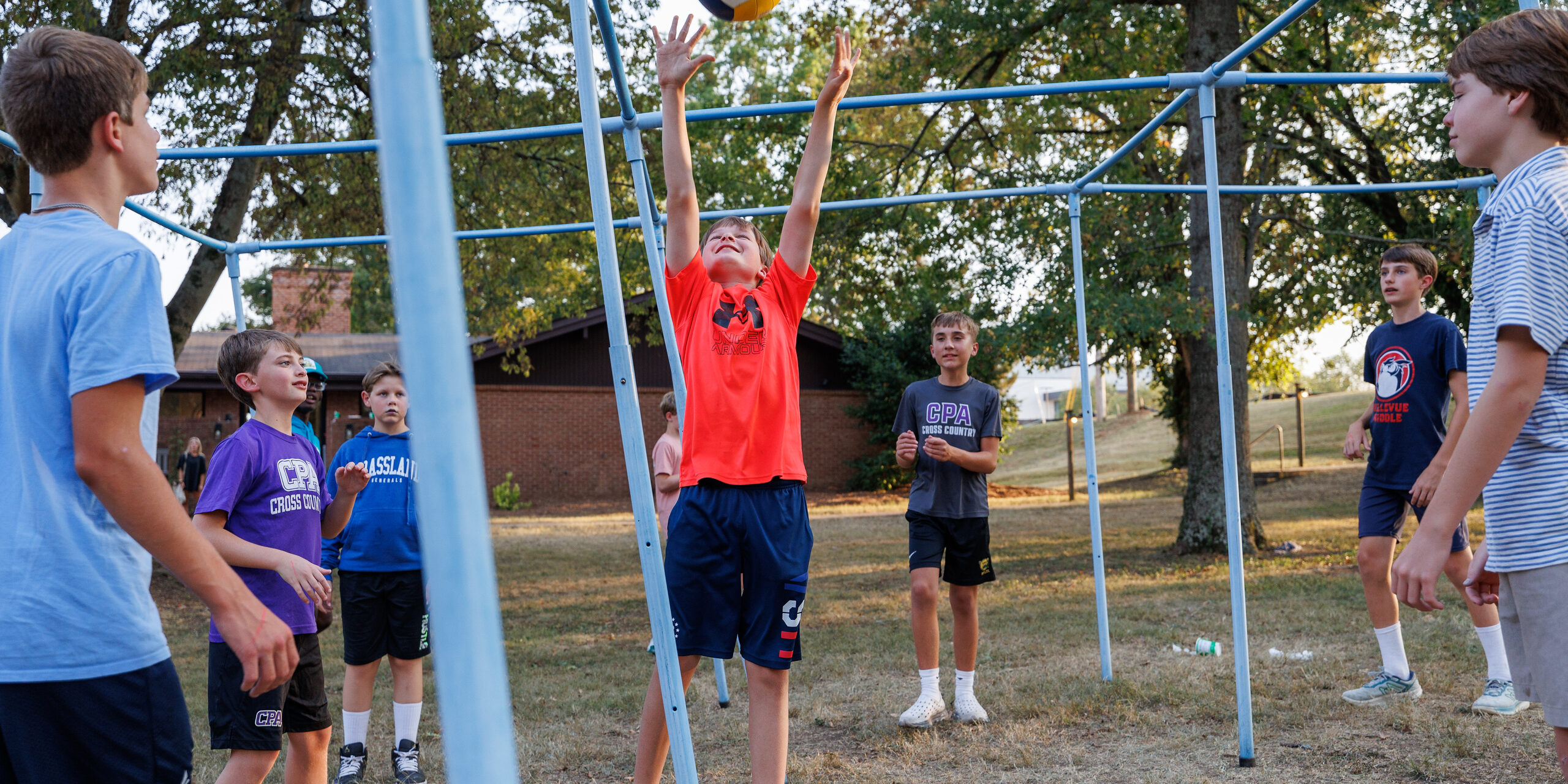 A group of boys play outdoor volleyball on a grassy field, with one boy in a red shirt jumping to hit the ball. Trees and a building are visible in the background. The scene is casual and energetic.