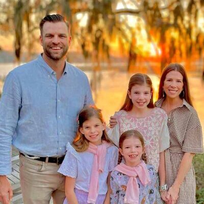 A smiling family of five poses outdoors near a lake at sunset. The parents stand behind their three young daughters, who wear pastel dresses and pink scarves. Trees and the glowing sun are in the background.
